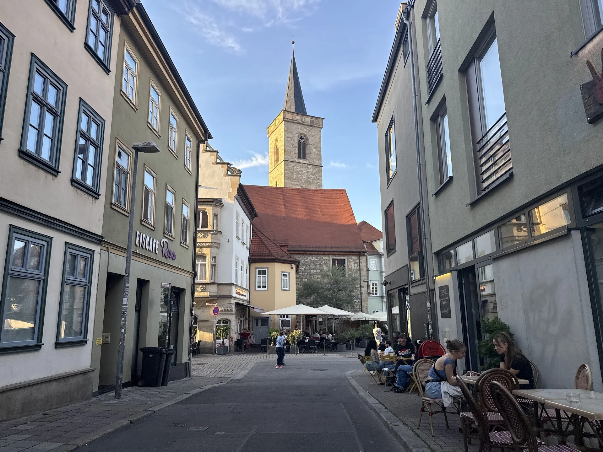 Street view of a lively outdoor café with people sitting at tables, surrounded by modern buildings, and a church steeple visible in the background under a clear sky.