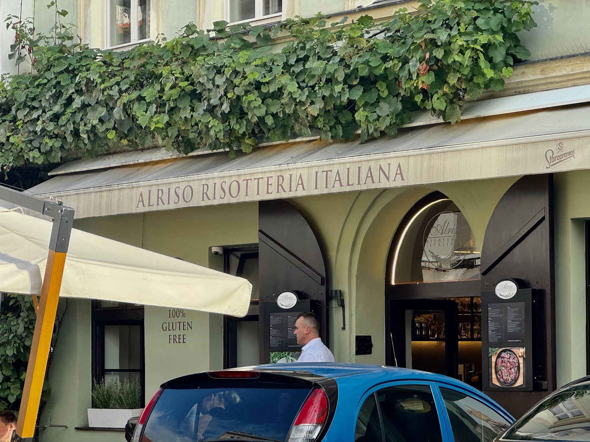 A storefront with a sign reading 'Alriso Risotteria Italiana' and a lush green vine on the upper part of the building. There are cars parked in front and a person standing outside.