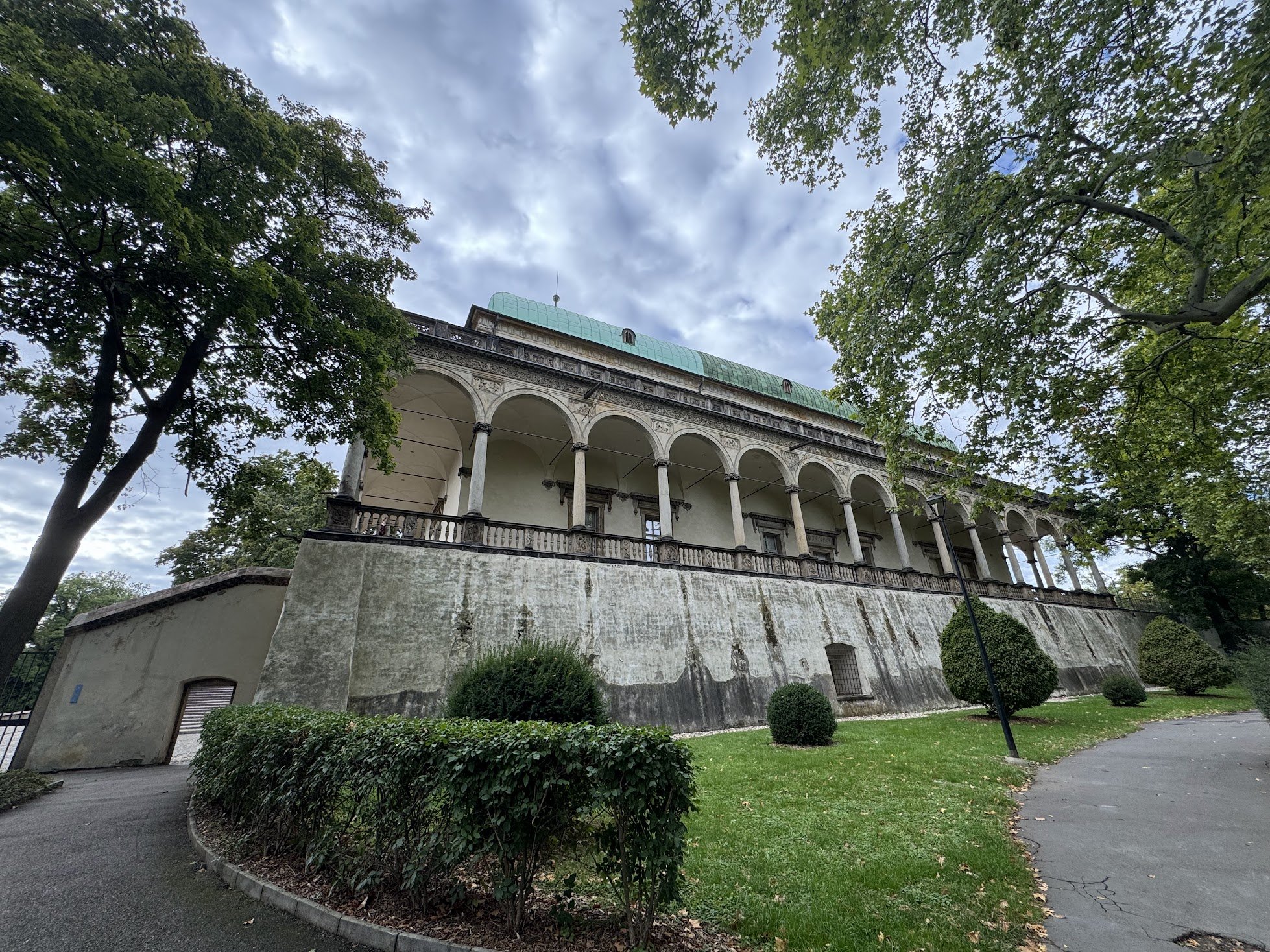 A historic building with a curved, green roof and tall arches on its upper level, surrounded by trees and a grassy area in a park-like setting during daytime.