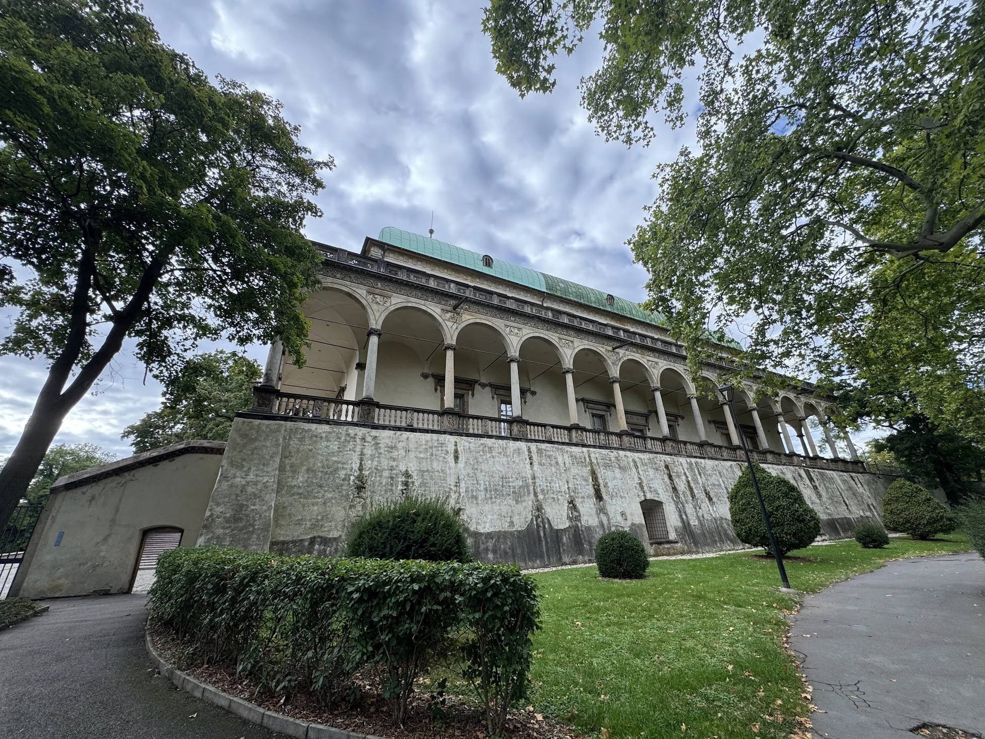 A historical building with arched windows and a green roof, surrounded by trees and a landscaped garden, under a cloudy sky.