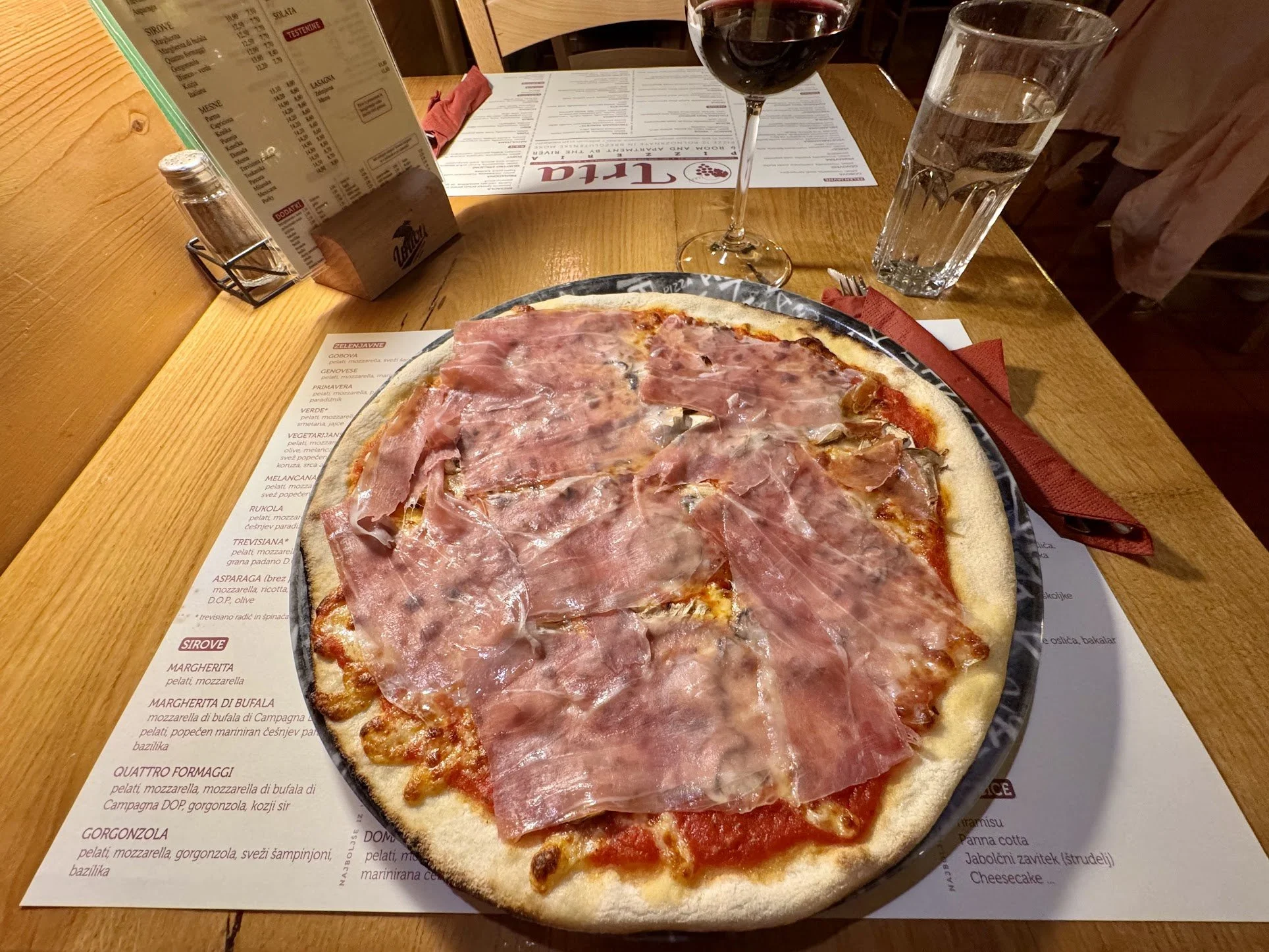 A pizza topped with slices of ham on a wooden table. Nearby are a glass of red wine, a glass of water, and a menu. The setting appears to be a restaurant.