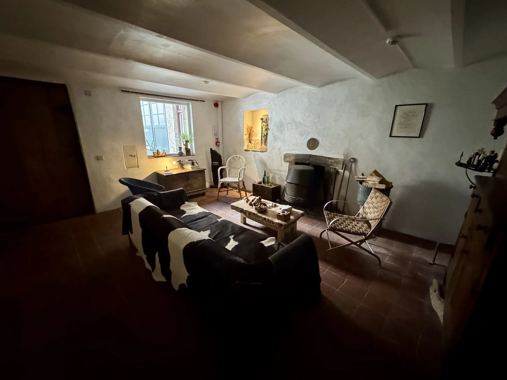 Cozy living room with beige textured walls, wood beam ceiling, window with blinds, and a mix of furniture including a black and white striped couch, a wooden coffee table, and various decorative items like bottles, a lamp, and framed art on the wall.