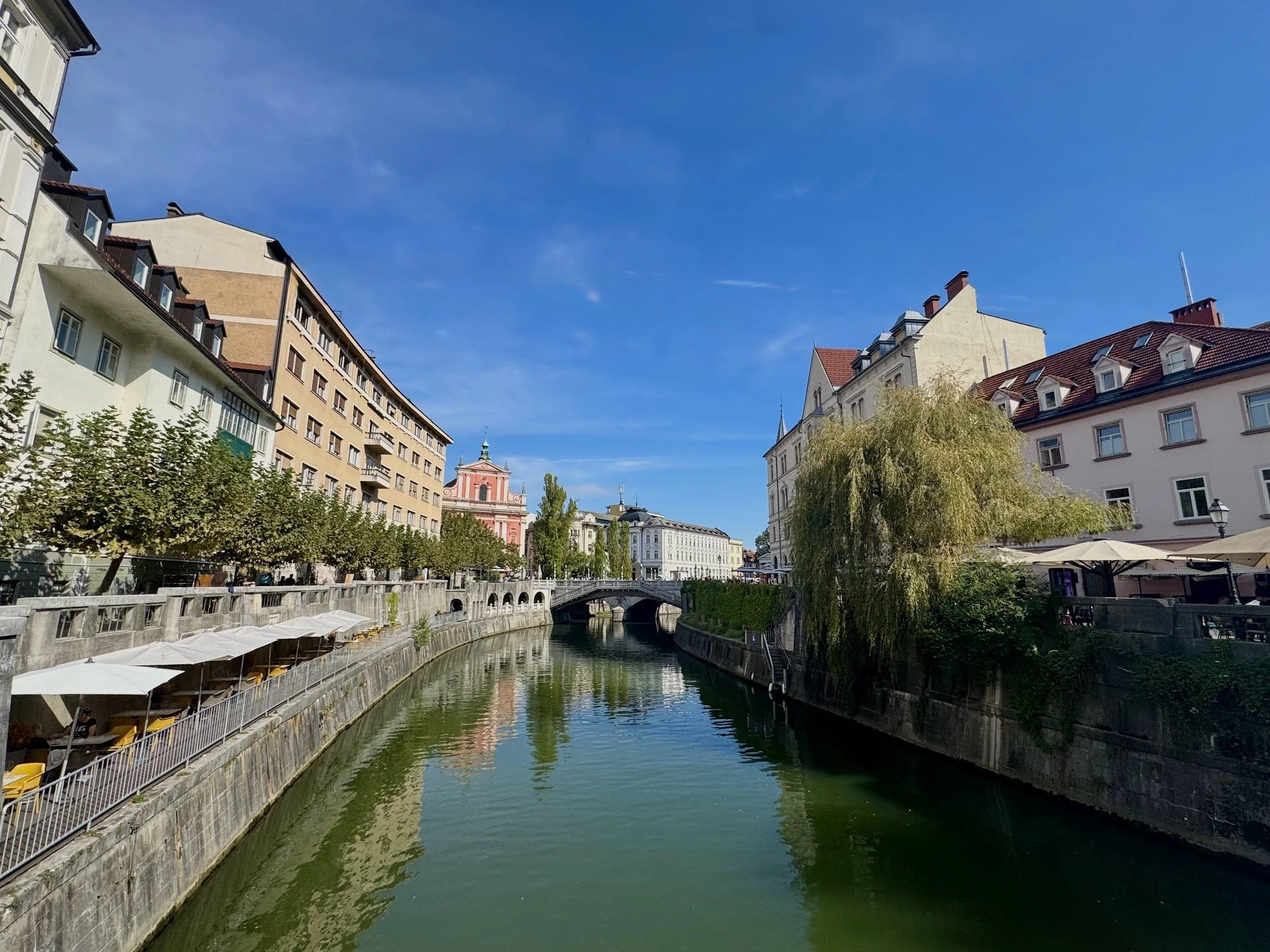 A peaceful cityscape featuring a canal lined with trees and outdoor seating, with multi-story buildings and a red church or historic building in the background under a clear blue sky.
