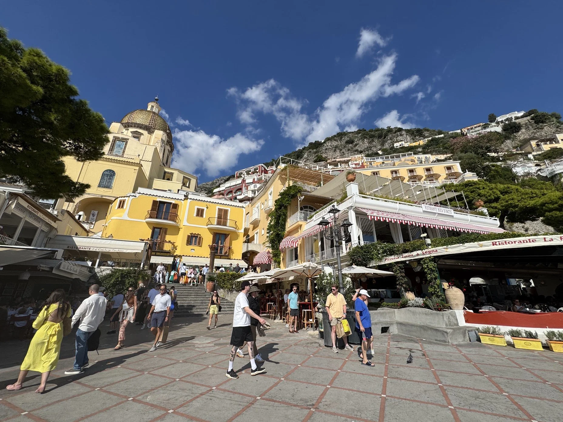 Tourists walking and sitting in a lively piazza with colorful buildings, shops, restaurants, greenery, and hillside houses against a blue sky with clouds.