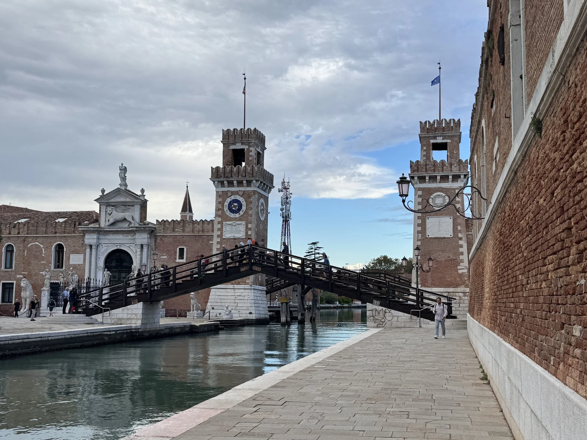 A historic brick building with two towers and flags, a bridge over a canal, and a statue on the roof, with people walking along the waterfront and cloudy sky above.