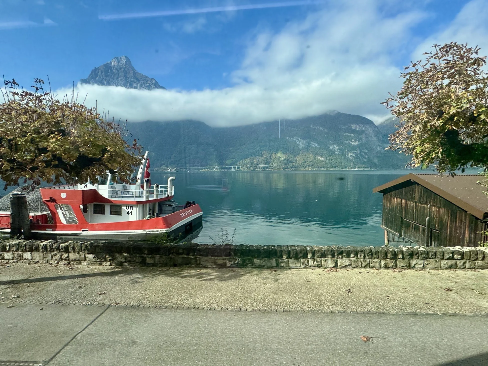 A red and white boat docked near a stone wall on a calm lake, with forested mountains in the background and partly cloudy sky.