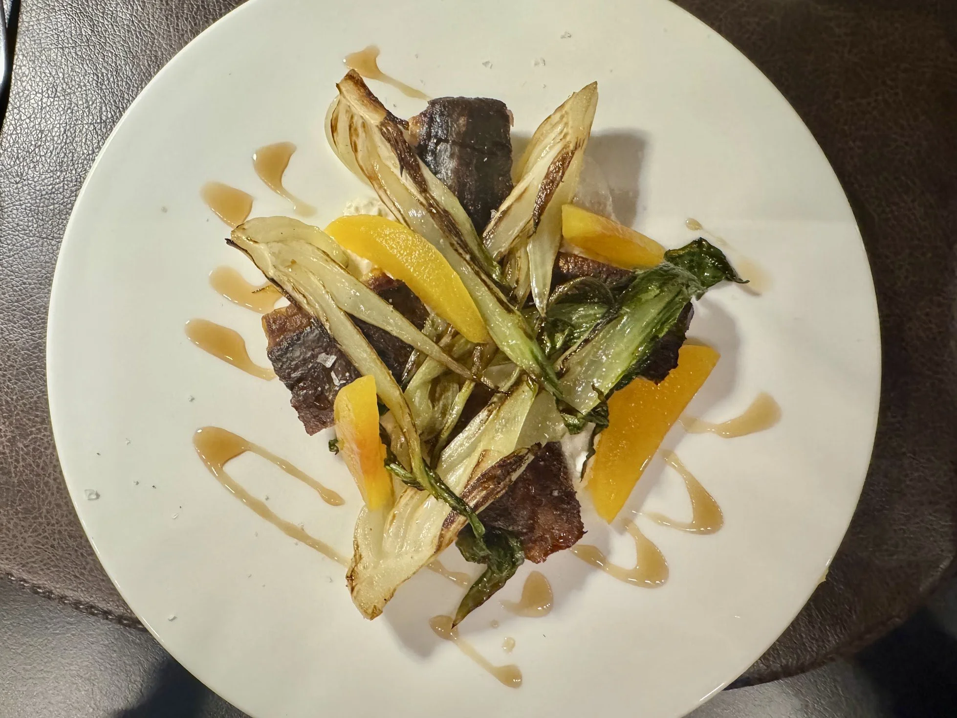 Seared steak with grilled vegetables including yellow squash, zucchini, and leafy greens, garnished with a drizzle of sauce on a white plate.