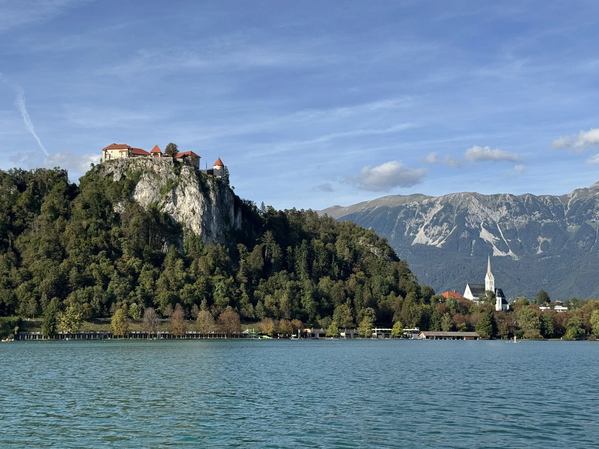 View of a mountain with a castle or historical building on top, a church with a steeple, trees along a hillside, and a large body of water in the foreground. The sky is partly cloudy.