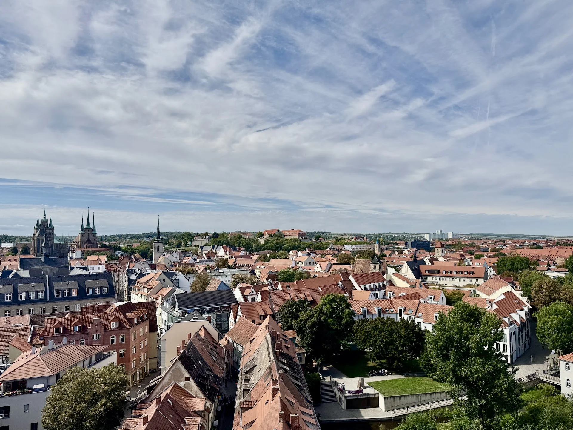 A cityscape viewed from above, showing buildings with red and gray rooftops, church spires, green trees, and distant hills under a partly cloudy sky.