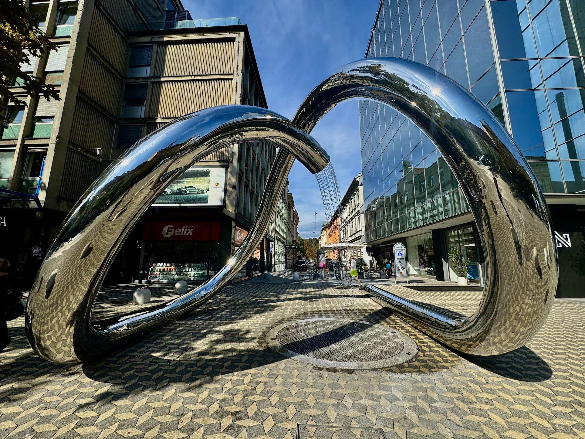 Large reflective metal sculpture resembling the number 20, situated on a patterned sidewalk in an urban setting. Buildings with glass windows and a street with pedestrians are visible in the background under a clear blue sky.