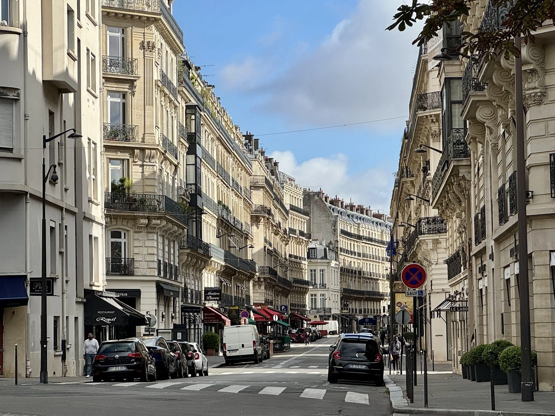 Parisian street scene with classic multi-story buildings, street cafes, parked cars, pedestrians, and blue sky.
