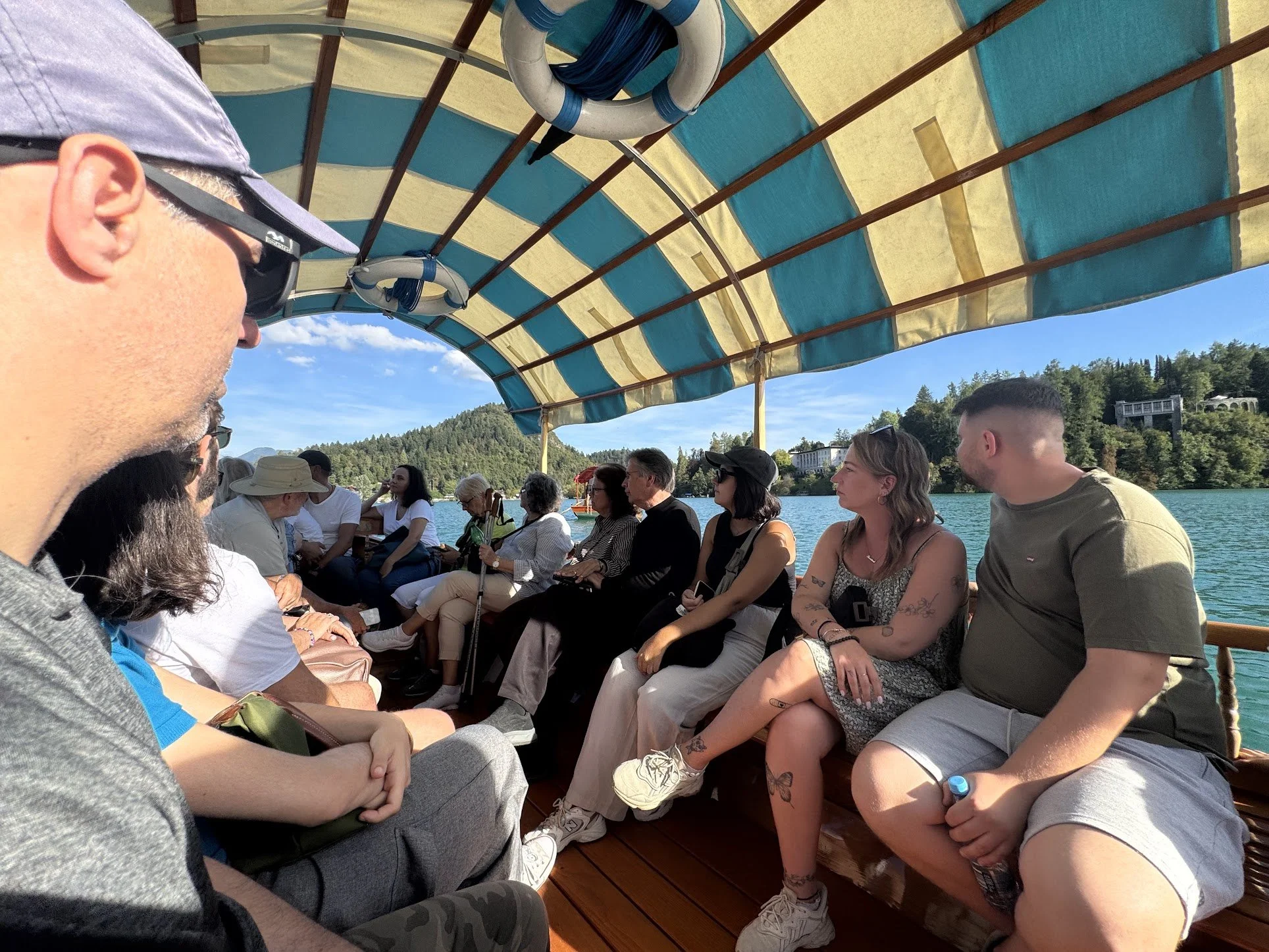 Group of people sitting on a boat under a canopy on a lake with hills and trees in the background.