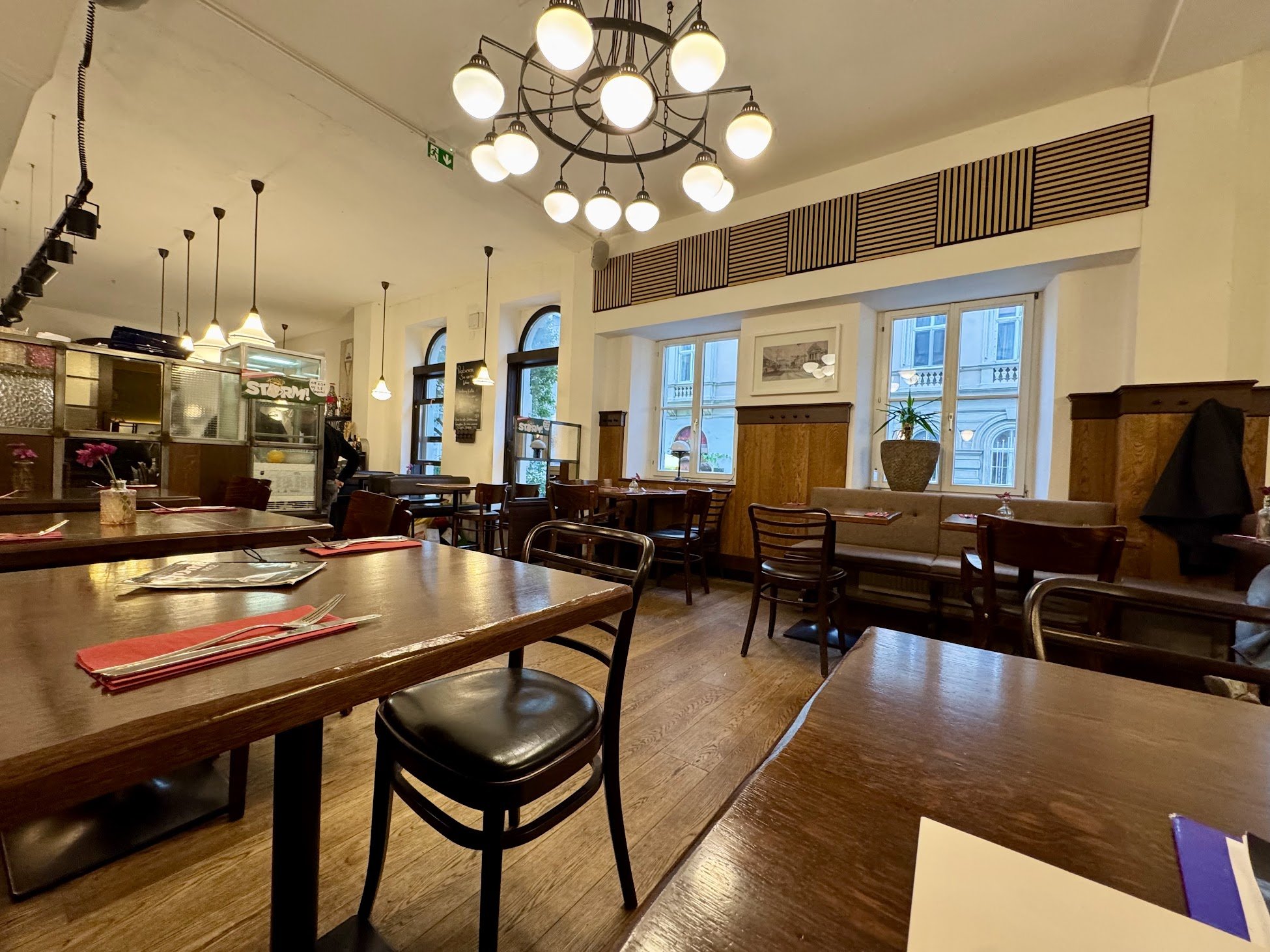 Interior of a cozy restaurant with wooden tables and chairs, large windows, hanging pendant lights, a chandelier, and decorative plants.