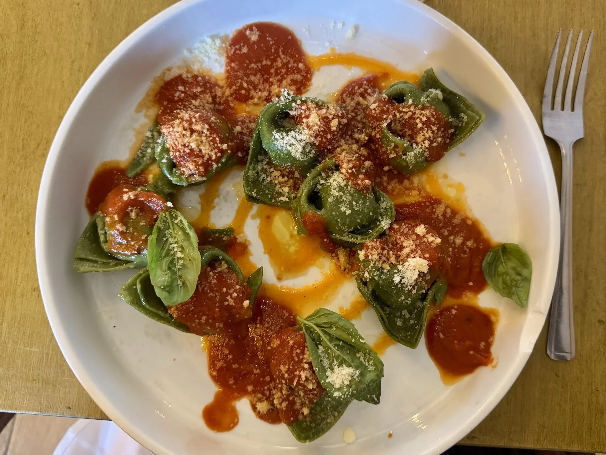 A white plate of cooked green bell pepper halves stuffed with cheese or sausage, topped with tomato sauce, grated cheese, and fresh basil leaves, on a wooden table with a fork nearby.