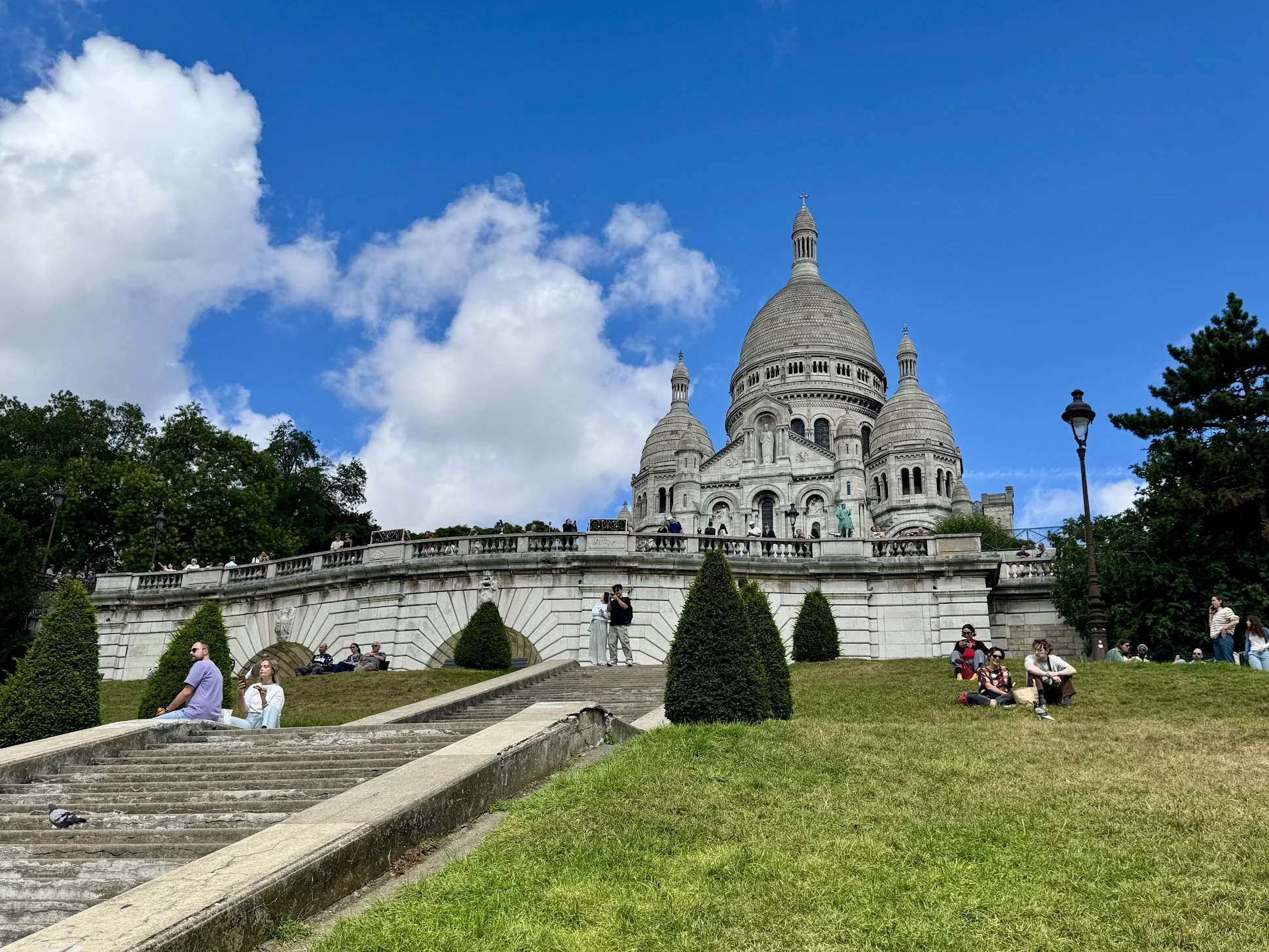 The Sacré-Cœur Basilica, a white Roman Catholic church with large domes, is seen in Montmartre, Paris. Several people are sitting and standing on the steps and grassy area in front of the church, with trees and a clear blue sky with clouds in the bac