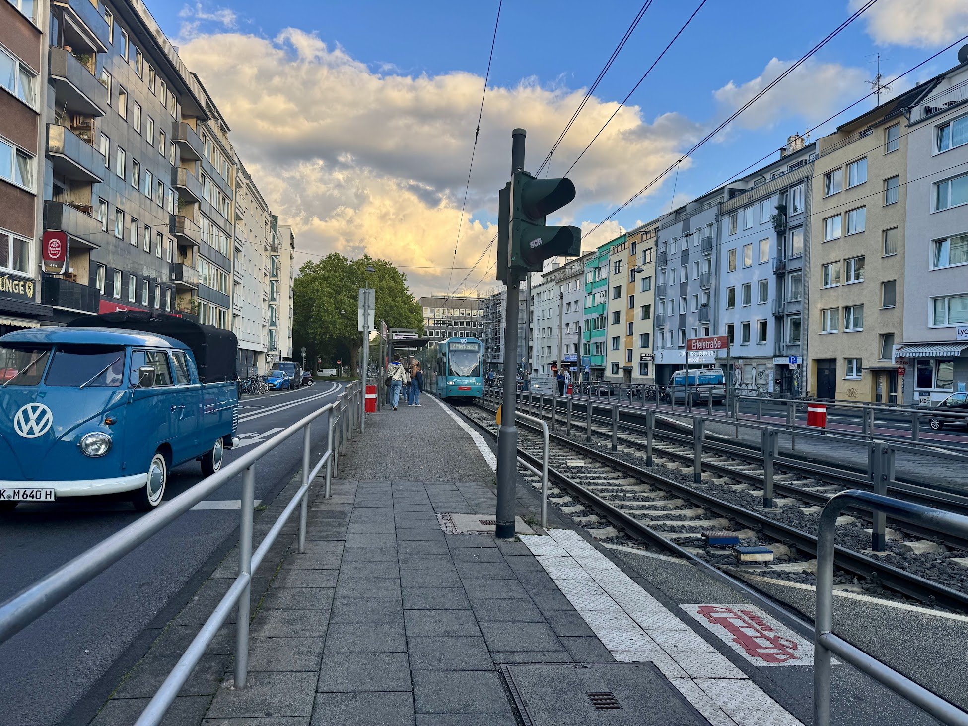 City street scene with a tram at a station, an old Volkswagen van parked on the left, multi-story residential buildings on both sides, and a partly cloudy sky overhead.