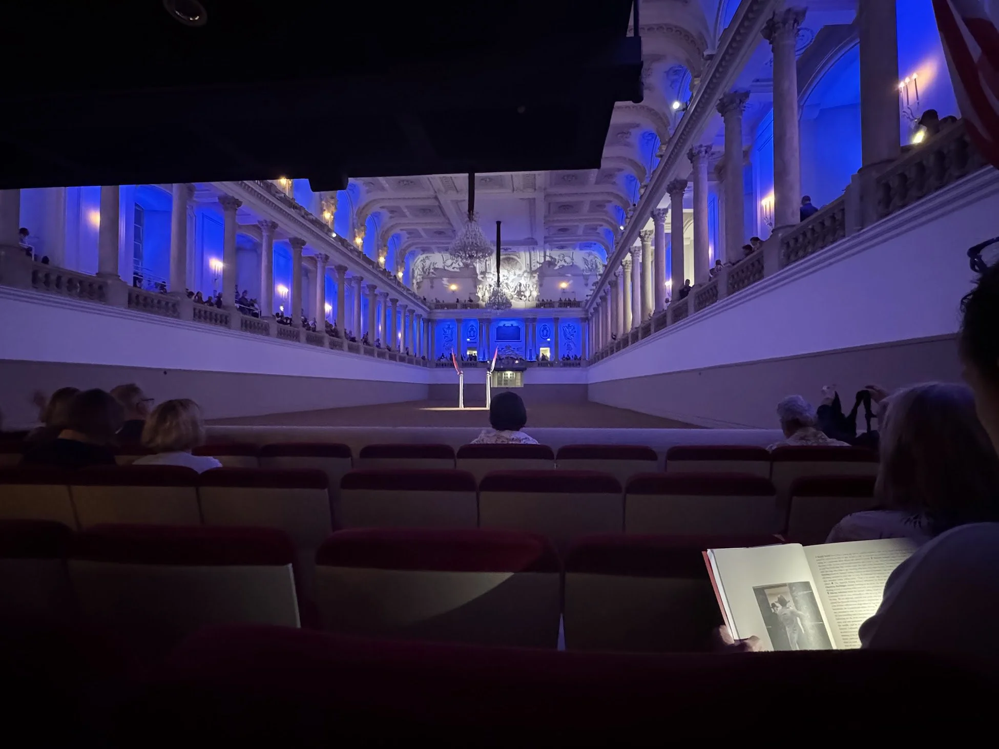 An indoor performance space with a high ornate ceiling, blue lighting, and a large chandelier. Audience members sit in rows of chairs watching a performance on a stage with two raised flags.