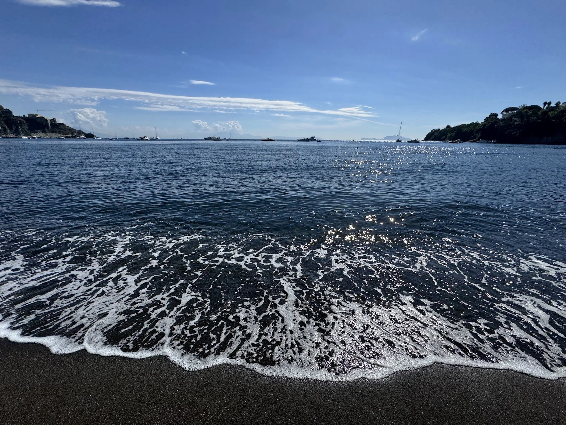 A view of the ocean near a sandy beach with boats anchored in the water and a rocky shoreline on the horizon under a partly cloudy sky.