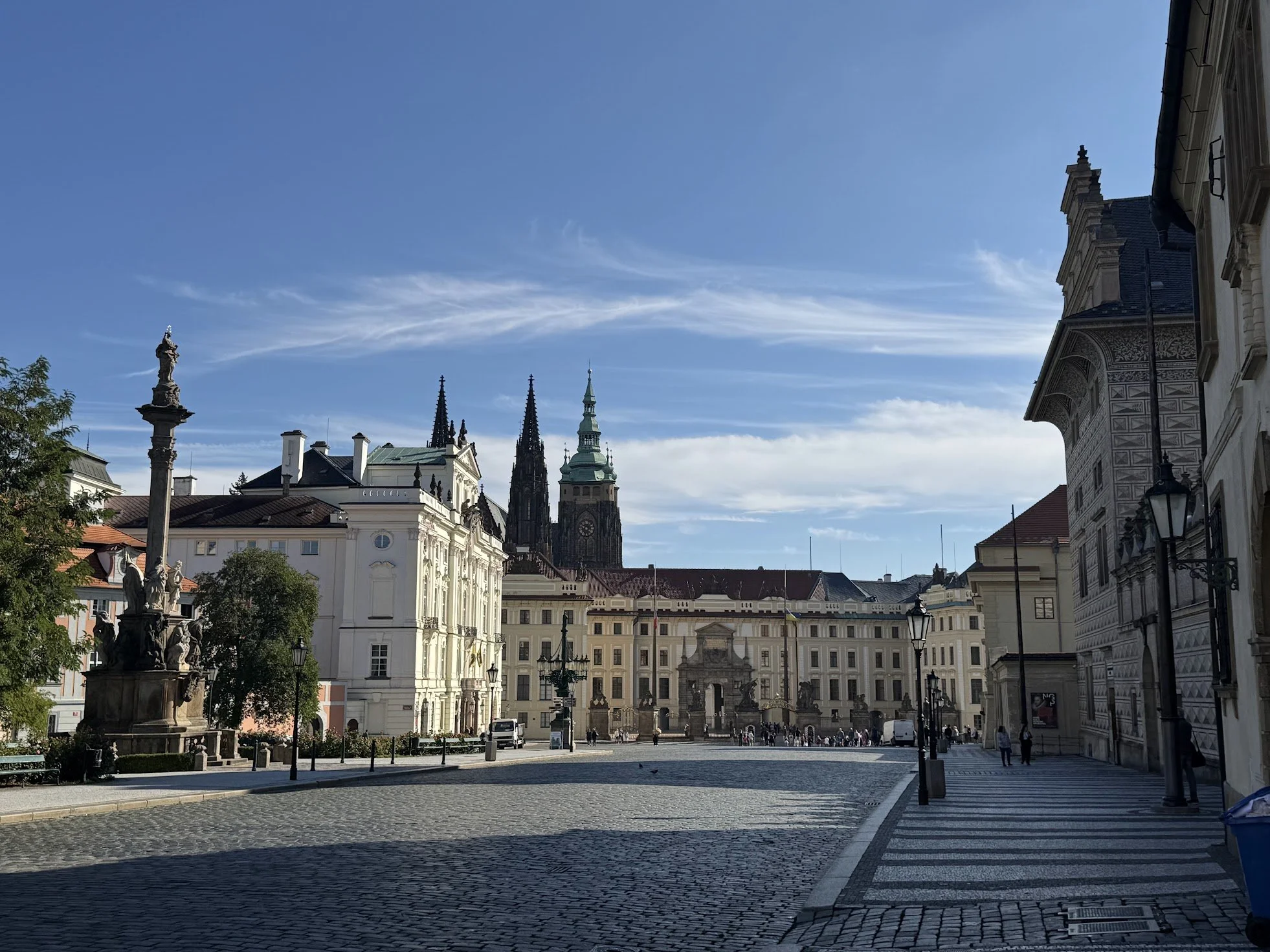 View of historic European city square with cobblestone pavement, ornate statues, and church spires under a blue sky with wispy clouds.