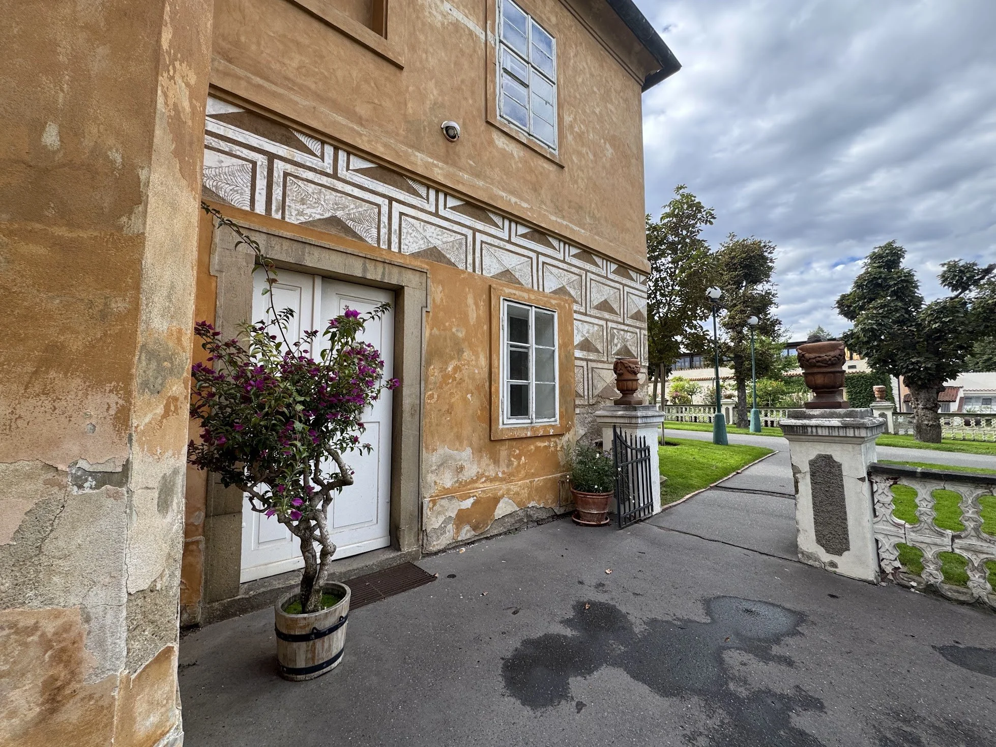 A building with a weathered, orange stucco exterior, white and brick patterned trim, and white framed windows, with potted plants and a pathway in a garden area with trees and decorative urns.