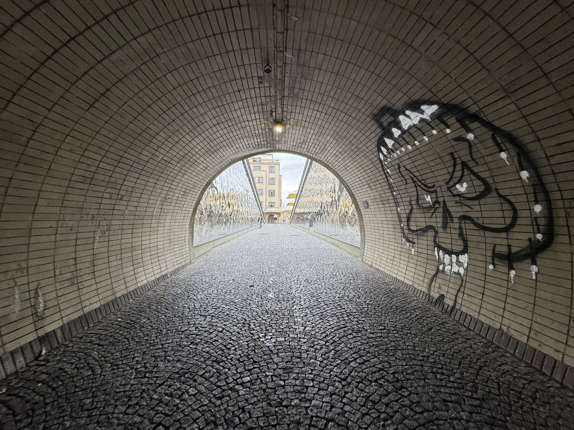 A tunnel with mosaic tiled walls and ceiling, graffiti on the right wall, cobblestone path inside, leading to a view of a building and cloudy sky outside.