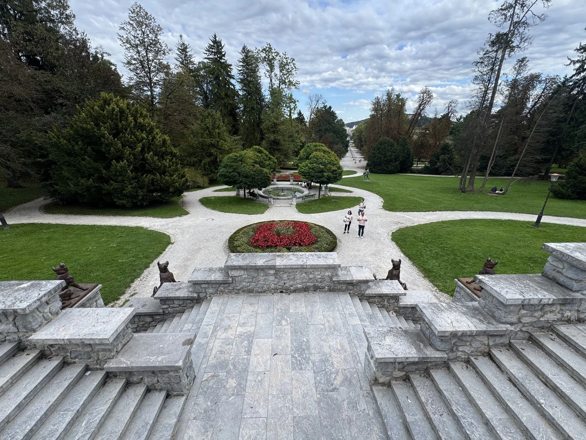 Stone steps lead to a garden with a circular fountain surrounded by trees and grass. People are walking and sitting in the park, with cloudy sky overhead.