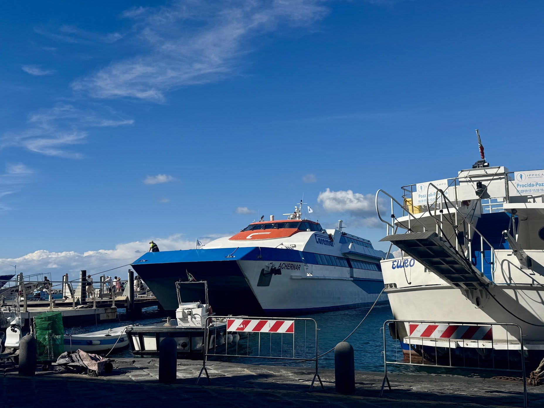 Boats docked at a harbor under a blue sky with some clouds, including one with a red and white color scheme and another with blue and white.