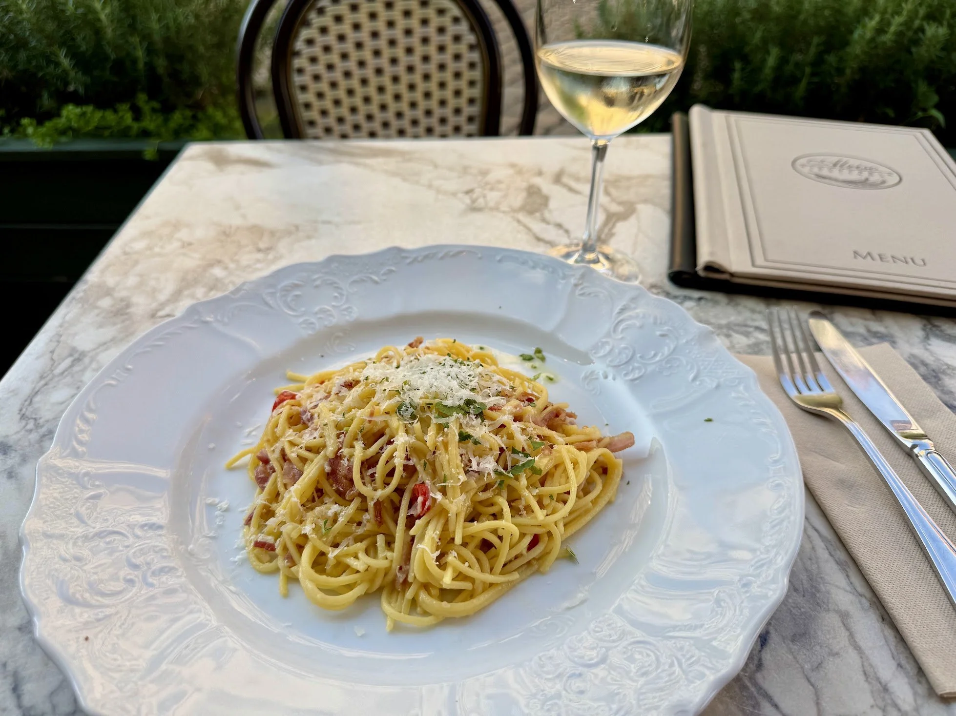 A plate of spaghetti carbonara topped with grated cheese and herbs on an ornate white plate. In the background, there is a glass of white wine, a menu, and a fork and knife on a marble table outdoors with greenery.