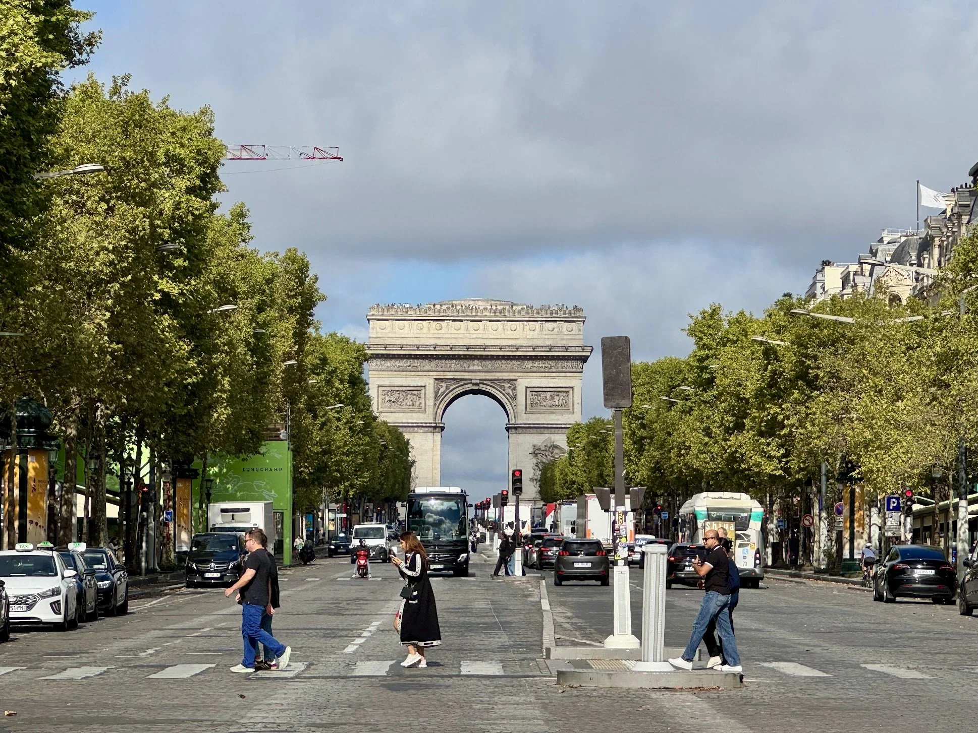 View of the Arc de Triomphe in Paris, France, seen through a busy city street with cars, pedestrians, trees, and a partly cloudy sky.
