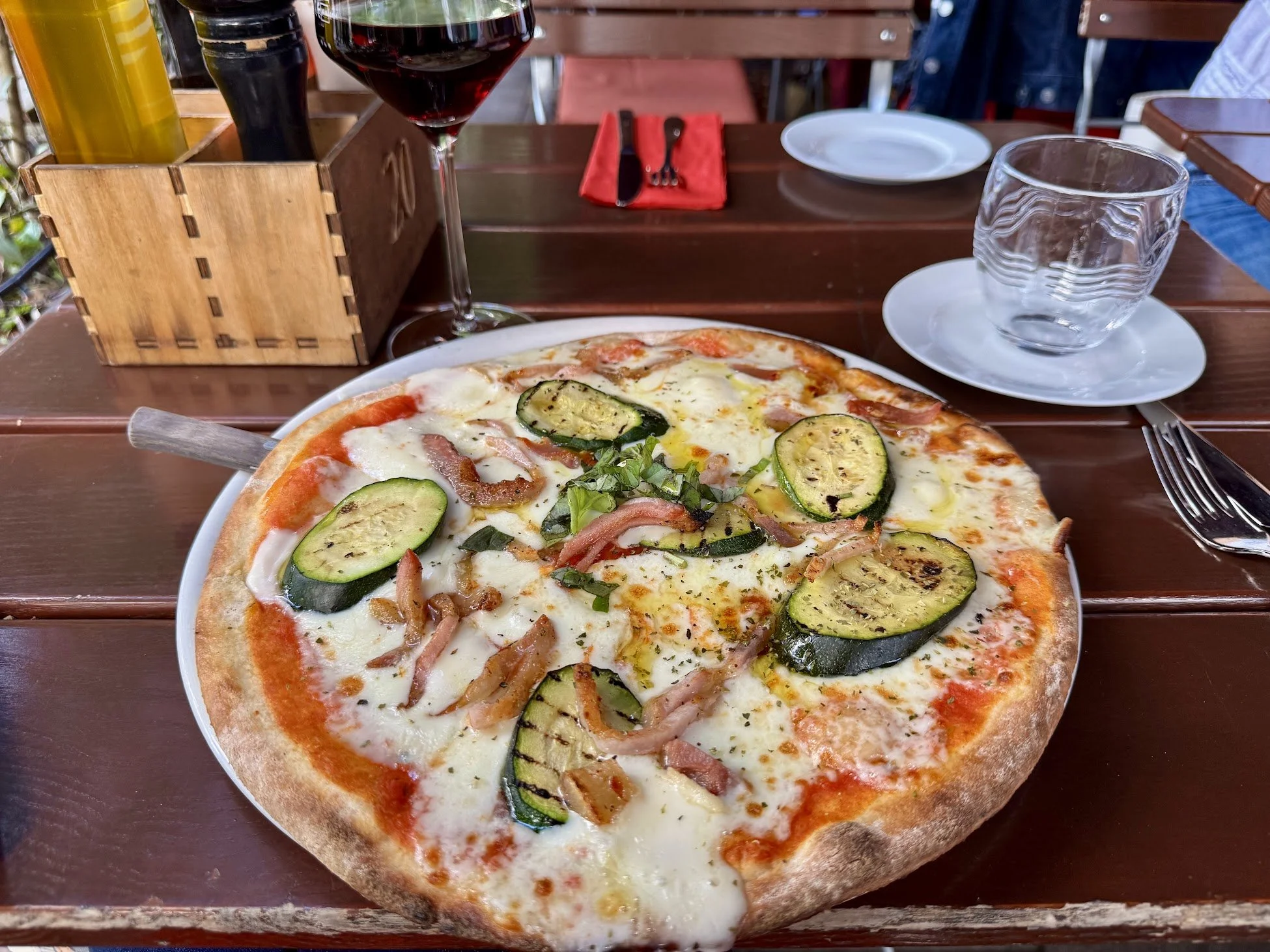 A pizza with cheese, zucchini, bacon, and herbs on a wooden table, with a glass of red wine, empty water glass, and utensils in the background.
