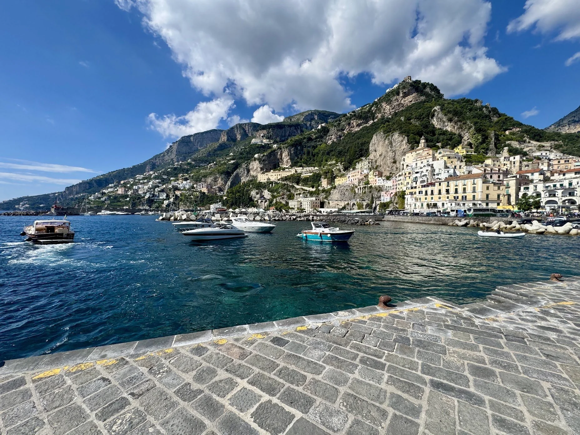 A coastal town with colorful buildings on a steep hillside beside a harbor filled with boats and yachts, with cloudy blue sky overhead and a stone promenade in the foreground.