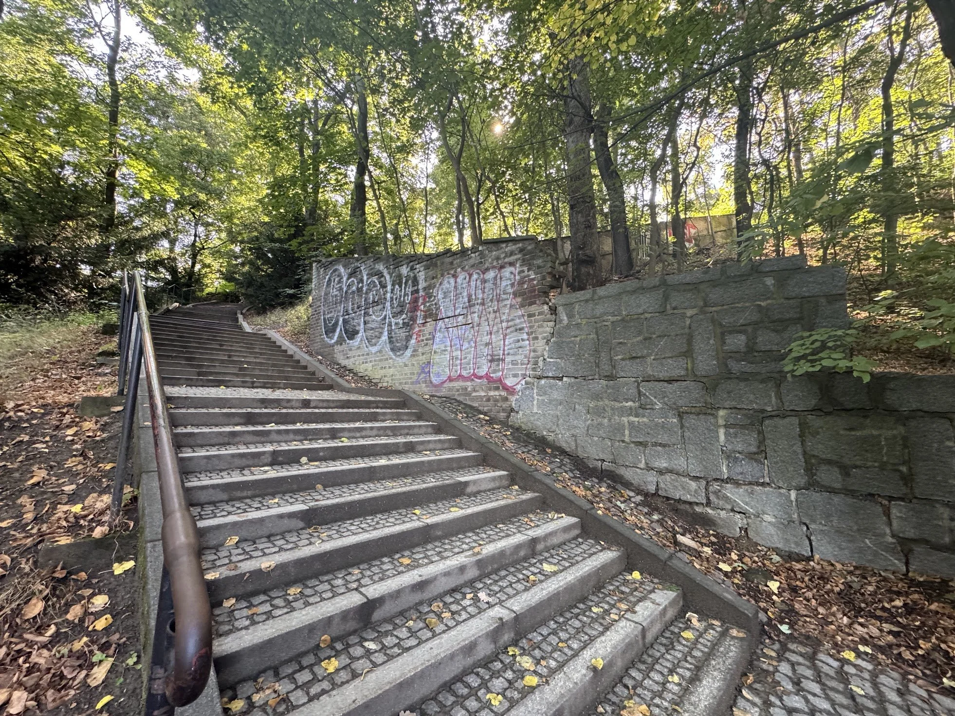 Outdoor stone staircase with a metal handrail, surrounded by trees with green foliage, some graffiti on a brick wall beside it, and fallen leaves on the ground.