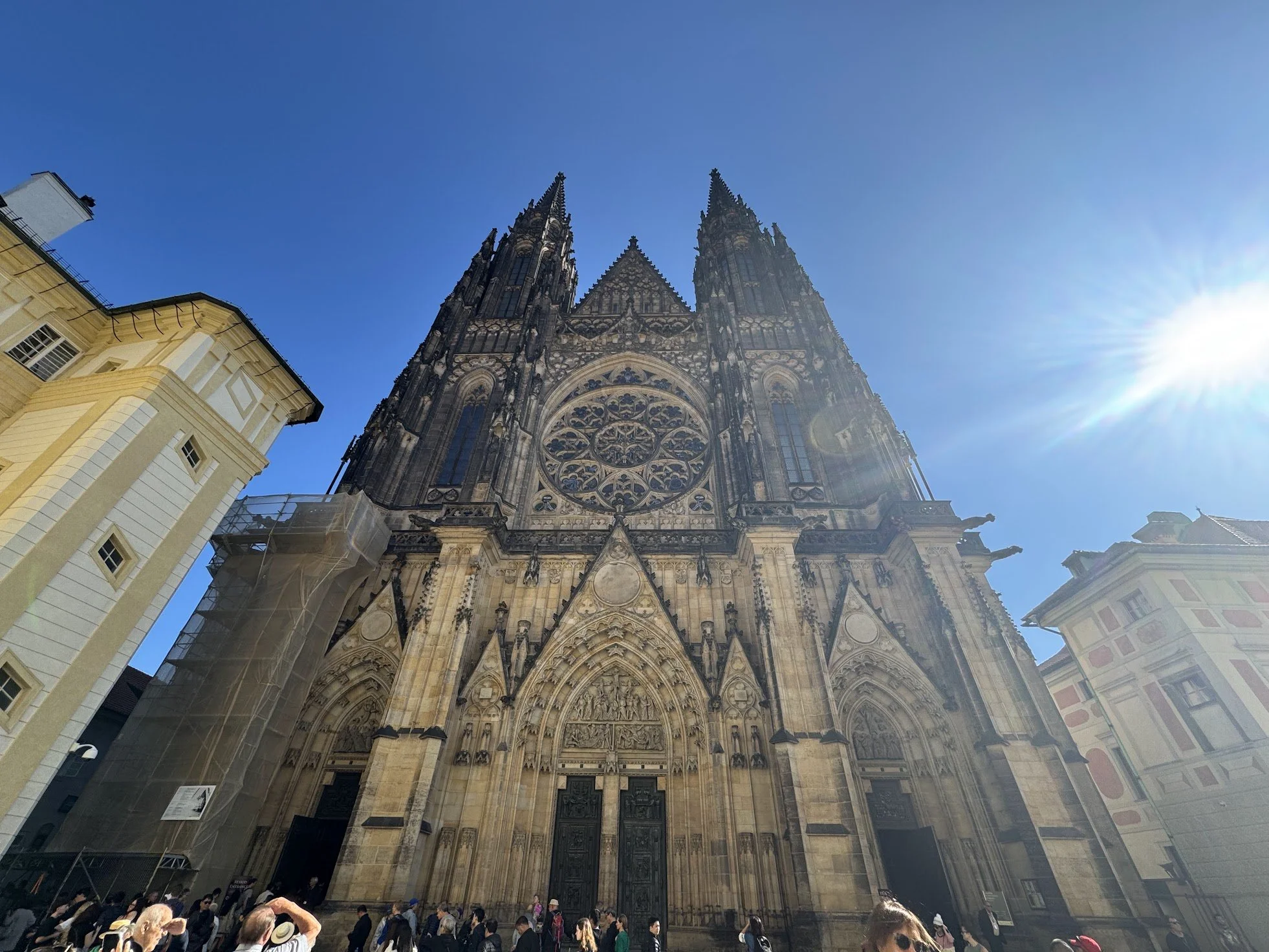 People gather outside a Gothic cathedral with two tall spires and ornate stone facade under a clear blue sky with bright sunshine.