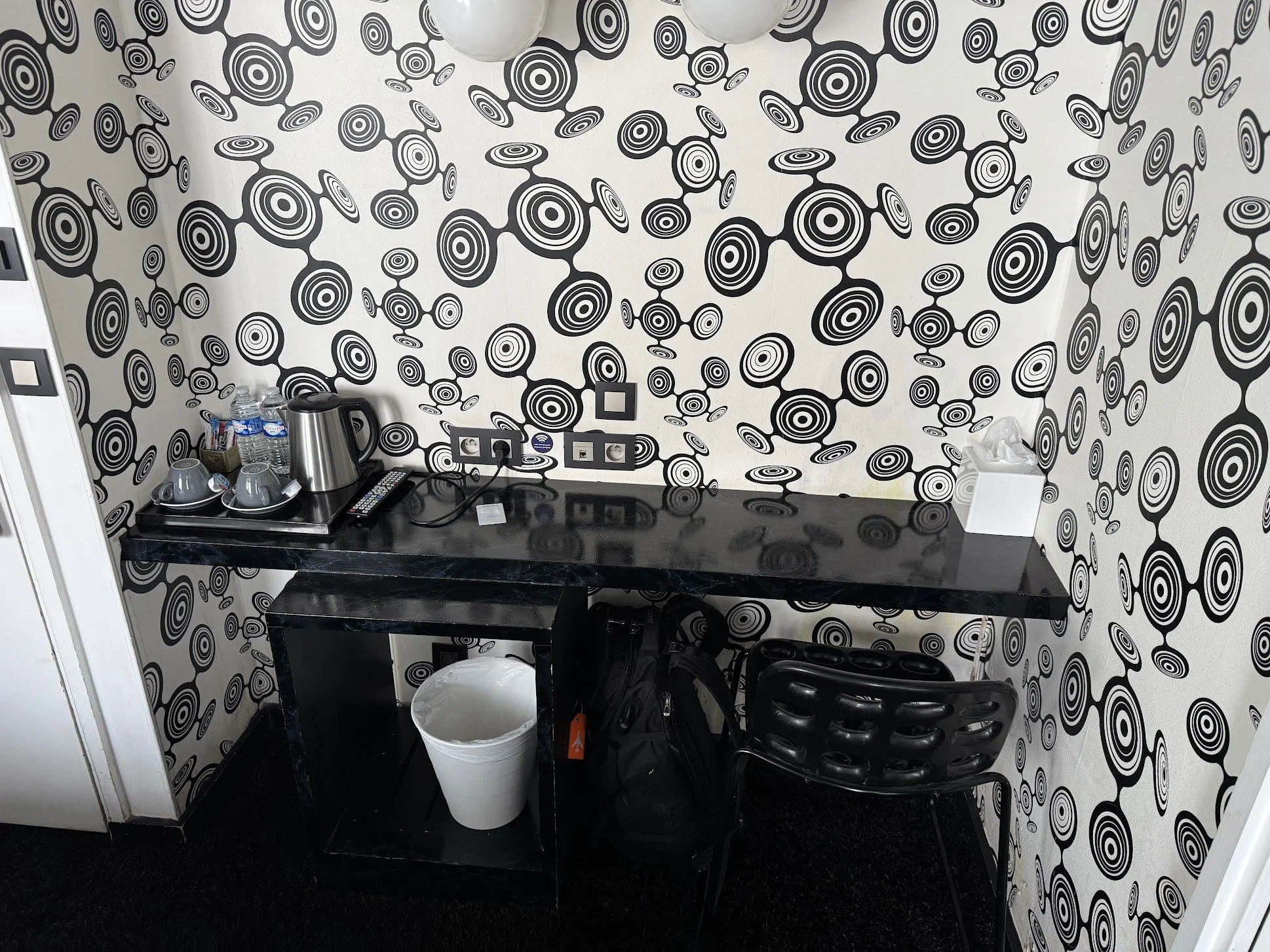 A black table against a wall with black and white circular wallpaper. On the table are bottled water, a metal electric kettle, mugs, a cordless phone, and some boxes. A white trash can is in front of the table, and a black backpack is underneath it. 