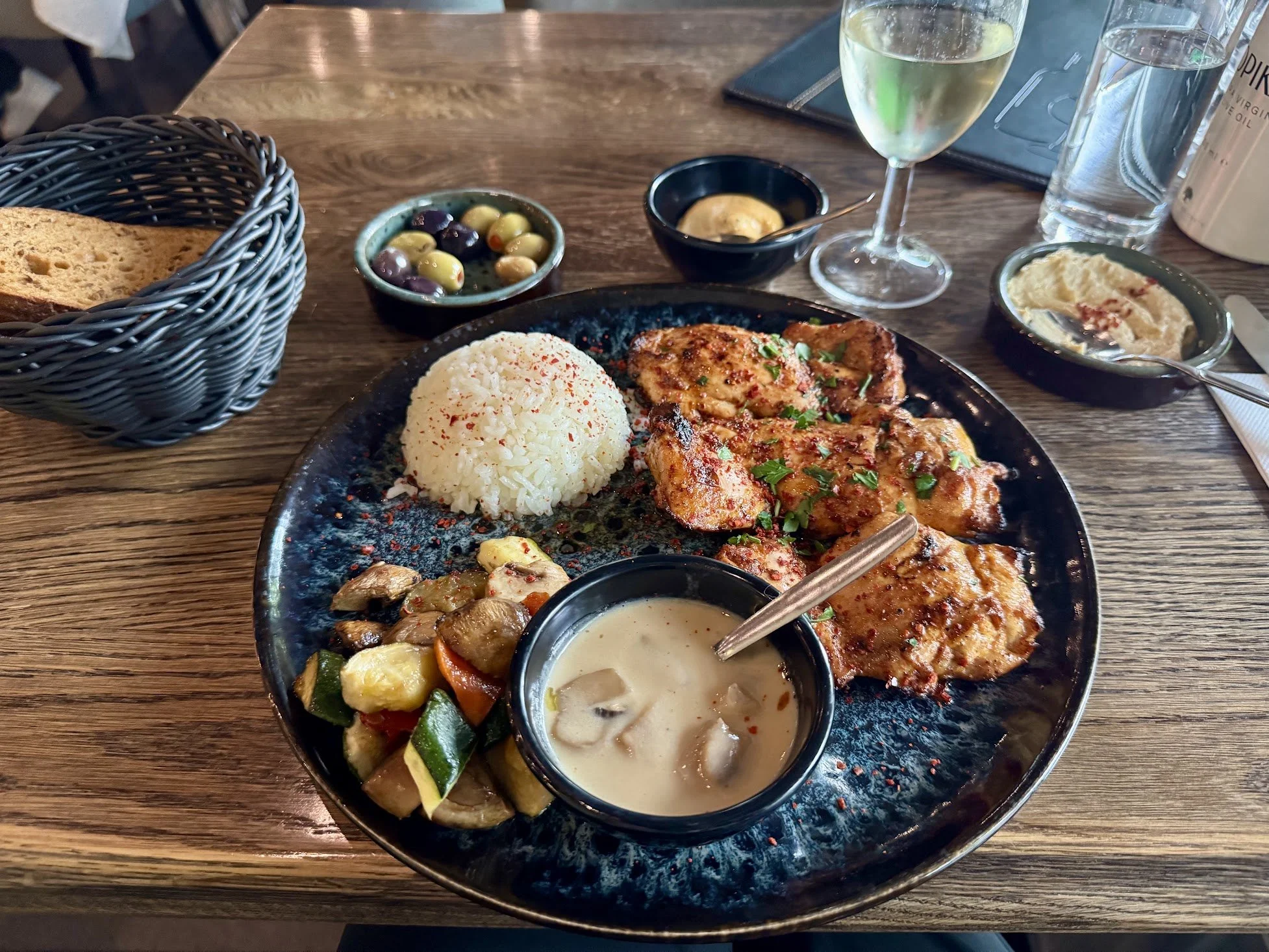 A plate of grilled chicken with rice, accompanied by roasted vegetables, cucumber salad, olives, hummus, and dipping sauces on a wooden table in a restaurant setting.