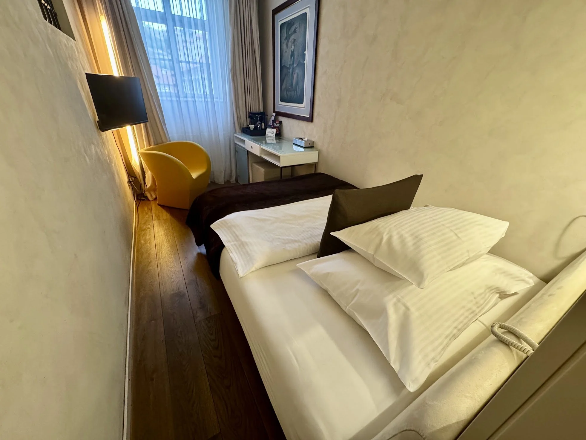 Hotel room with a bed, pillows, a small desk with a chair, a flat-screen TV, and a large window with curtains. Wall art hangs above the desk.