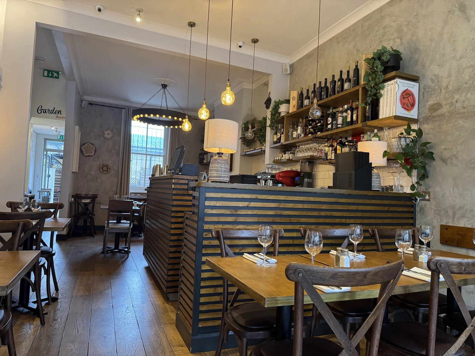 Inside a cozy restaurant with wooden furniture, a bar with bottles of alcohol on shelves, hanging pendant lights, and a large window.