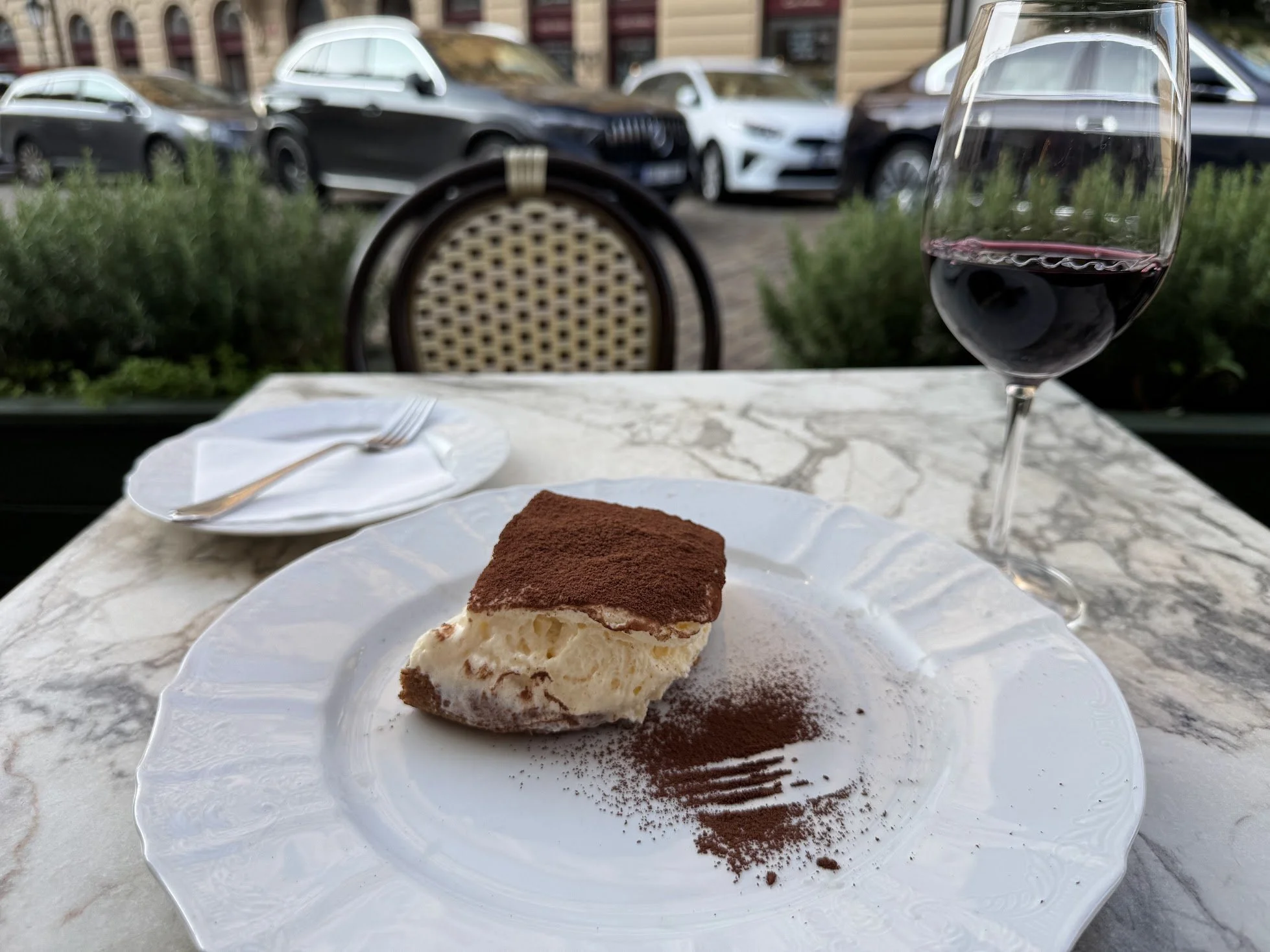 Slice of tiramisu dessert on a white plate, with a dusting of cocoa powder, on a marble table outdoors. In the background, a glass of red wine, a fork, and a napkin are visible, with cars parked outside.