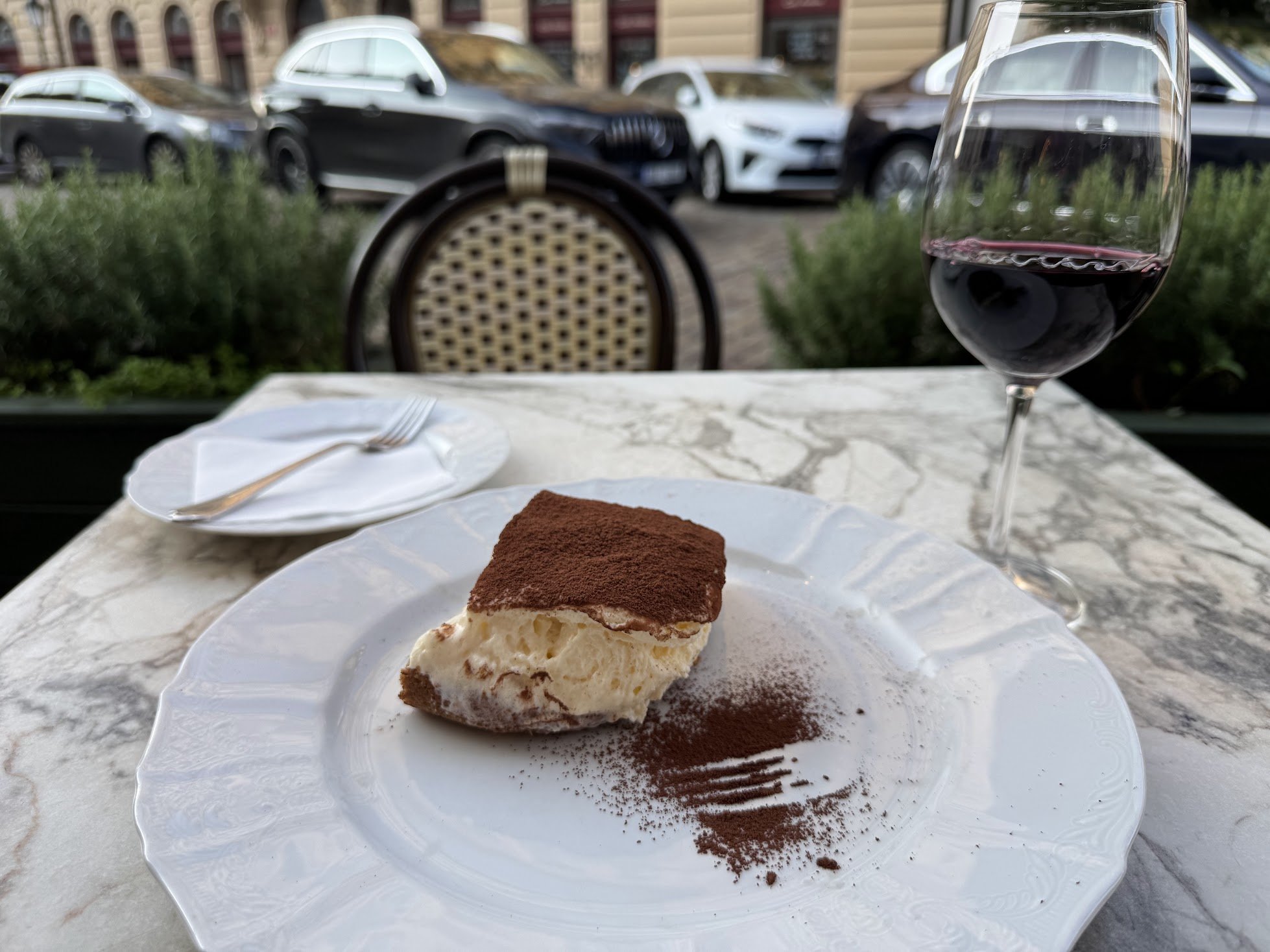 A slice of tiramisu on a decorative white plate with cocoa powder sprinkled on and around it. In the background, there is a glass of red wine, a fork and a napkin on a marble table. The scene is set outdoors with parked cars and a building visible in