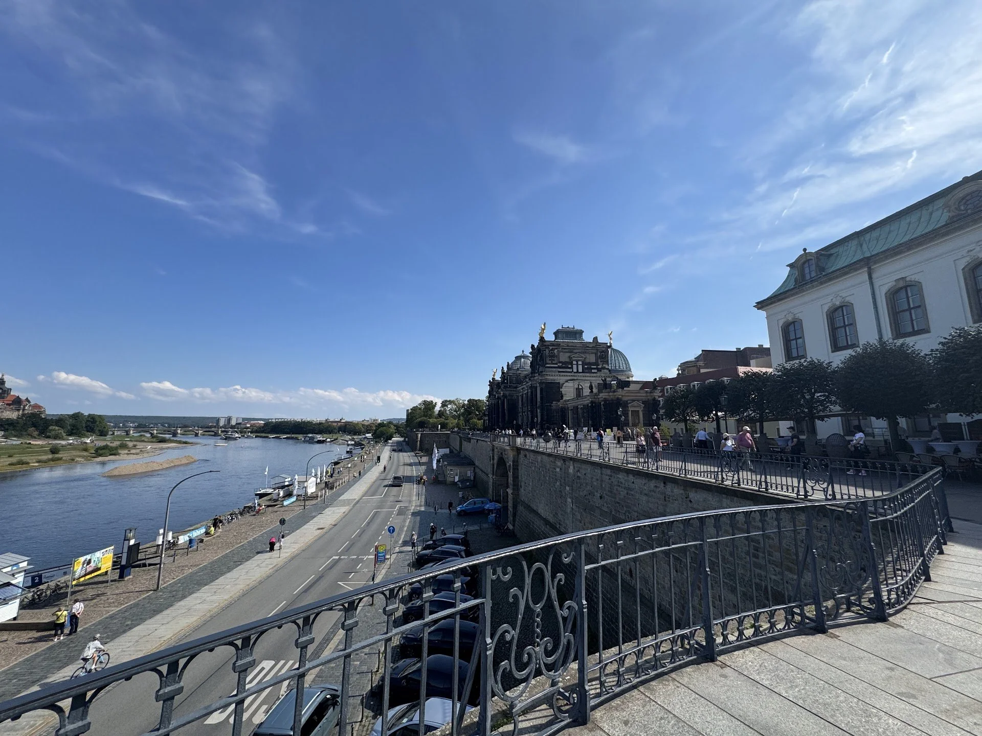 View of a river with a footpath and road along the bank, historic buildings on a hillside, and a clear blue sky with some clouds.