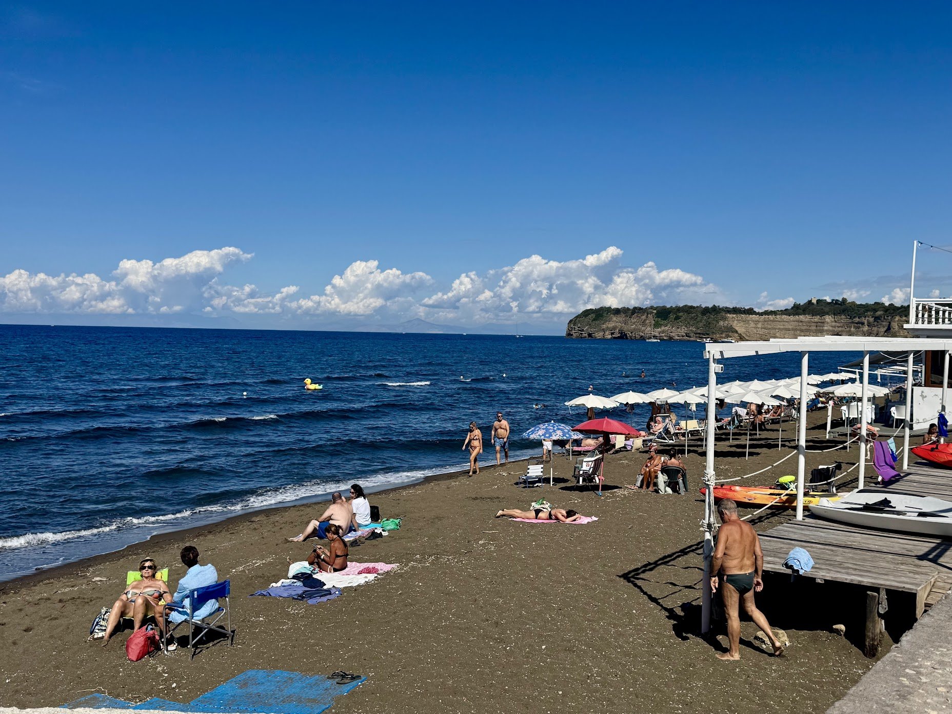 A crowded beach with people sunbathing on chairs and towels, some under umbrellas. The ocean is in the background with a rocky headland and a blue sky with white clouds.