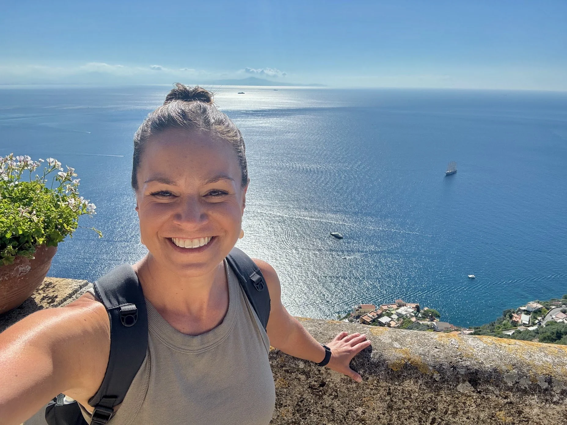 Smiling woman taking a selfie on a cliff with a view of the ocean, boats, and a coastline in the background.