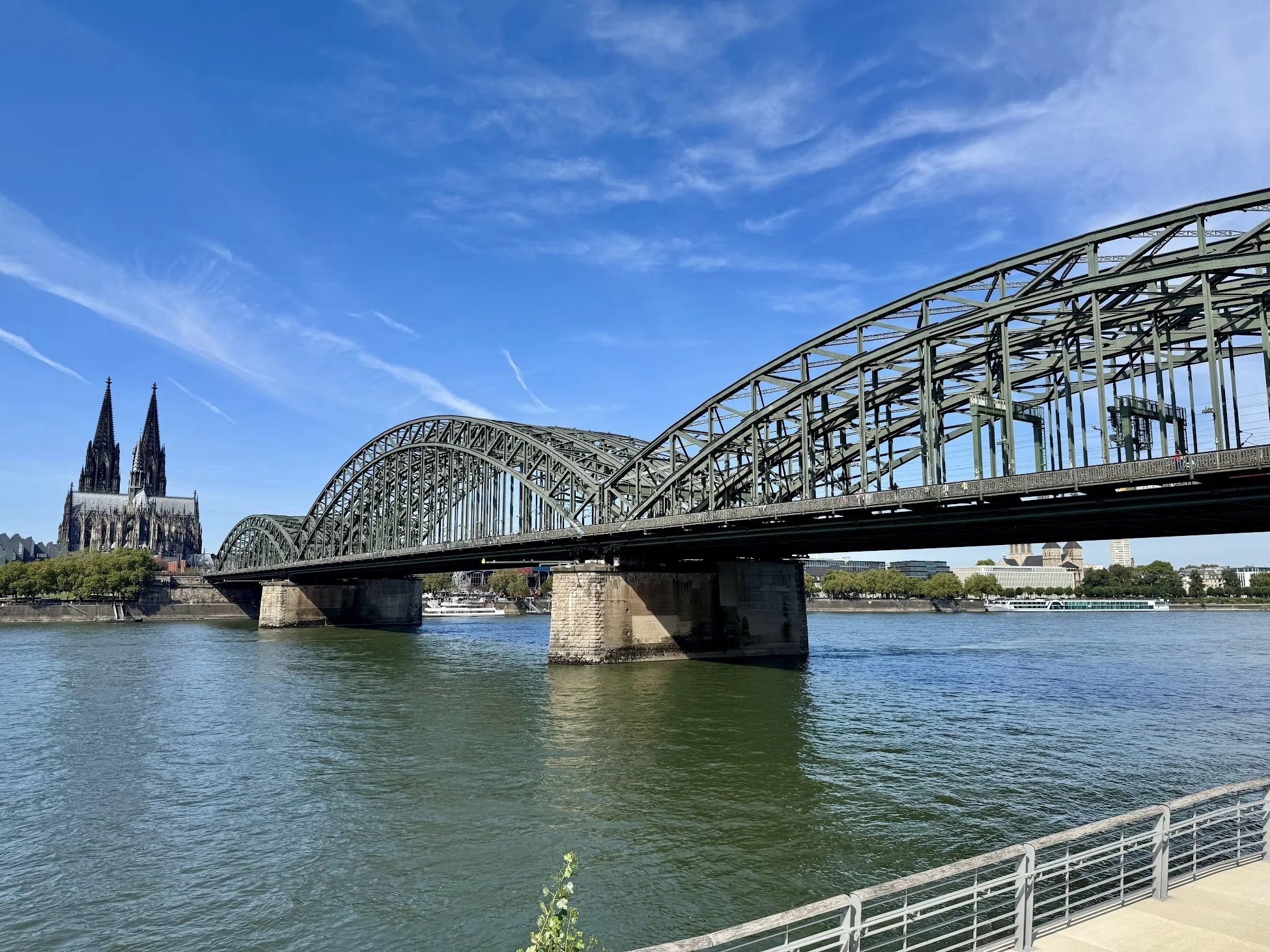 View of a metal bridge over a river with a Gothic cathedral in the background and clear blue sky.