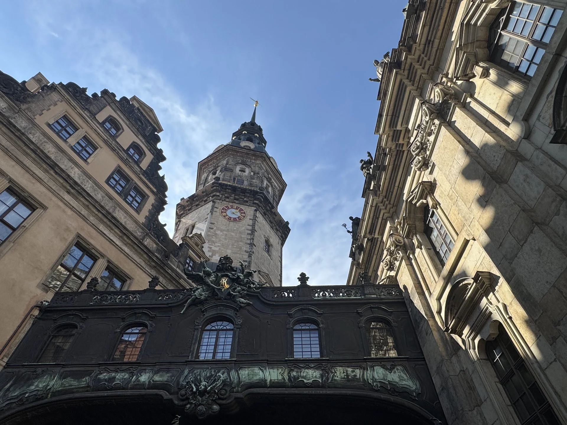 Looking up at historic European-style buildings with ornate facades and sculptures, including a clock tower with a spire, set against a blue sky with some clouds.