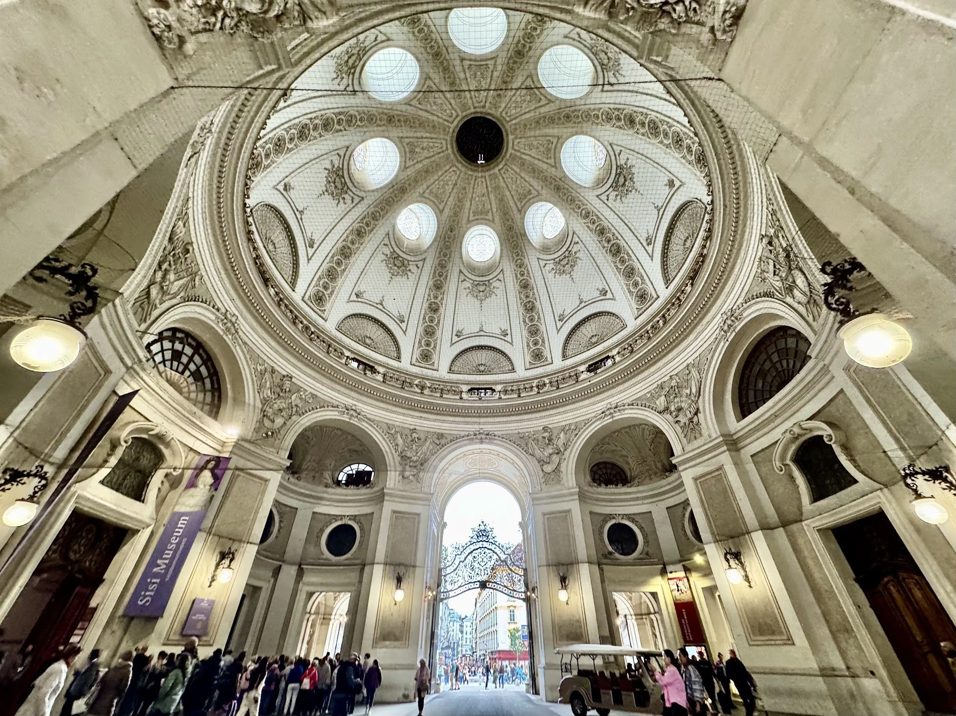 Inside a grand, ornate building with high vaulted ceiling, detailed decorative moldings, and large circular skylights. The interior features arches, sculptures, and light fixtures. People are walking and gathering inside, with shop signs visible at t