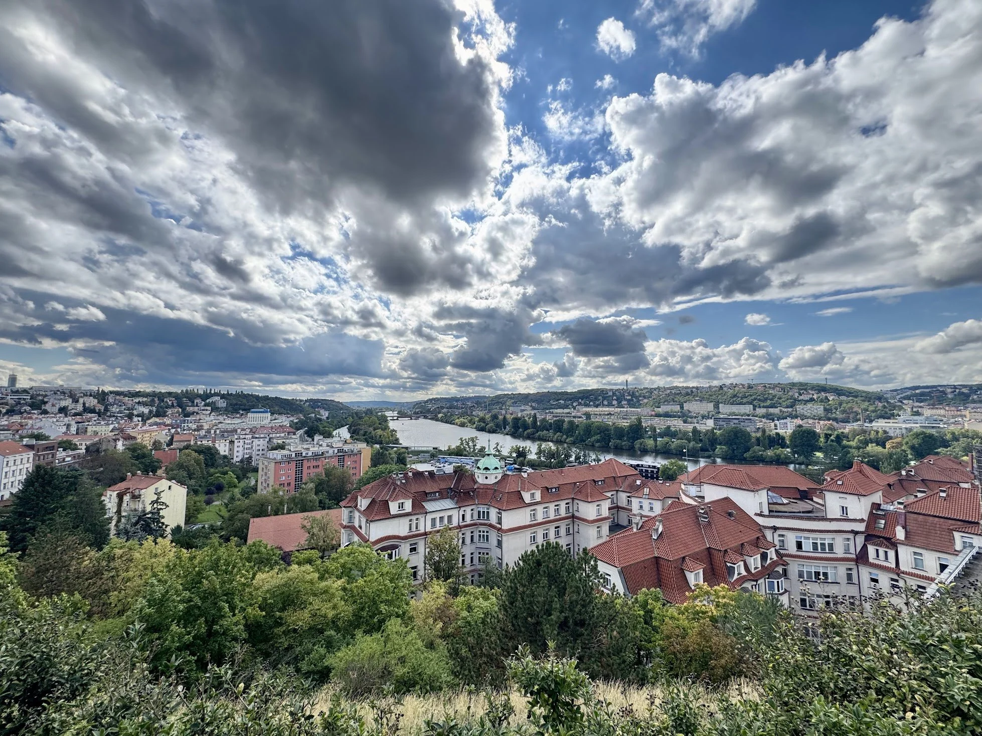 Cityscape view of a river running through the city, surrounded by buildings with red rooftops, greenery, and a partly cloudy sky with dark and white clouds.