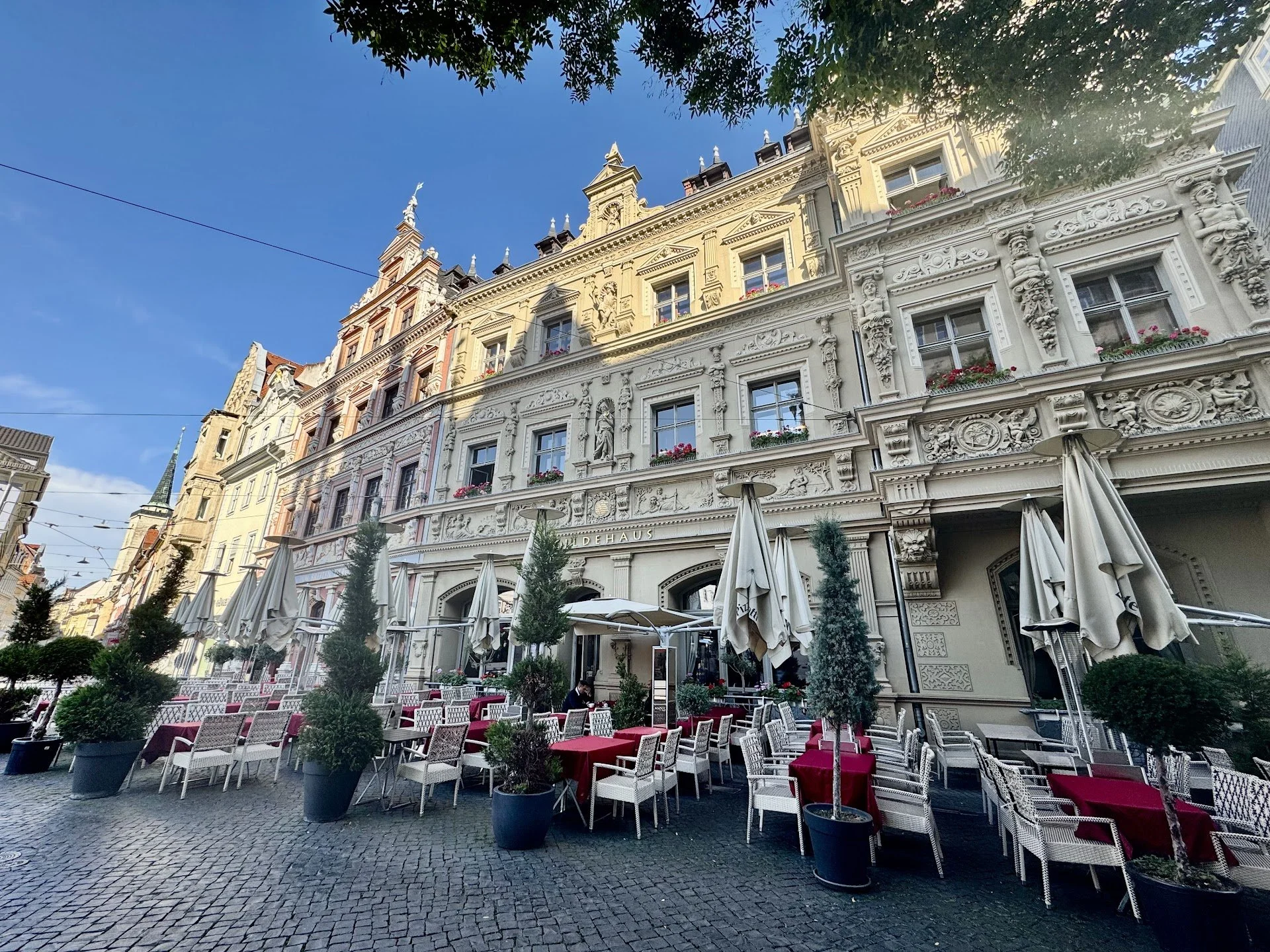 Outdoor seating area with white chairs and red-tablecloth-covered tables in front of an ornate historic building with sculptures and floral window boxes. Potted trees line the patio under a clear blue sky.
