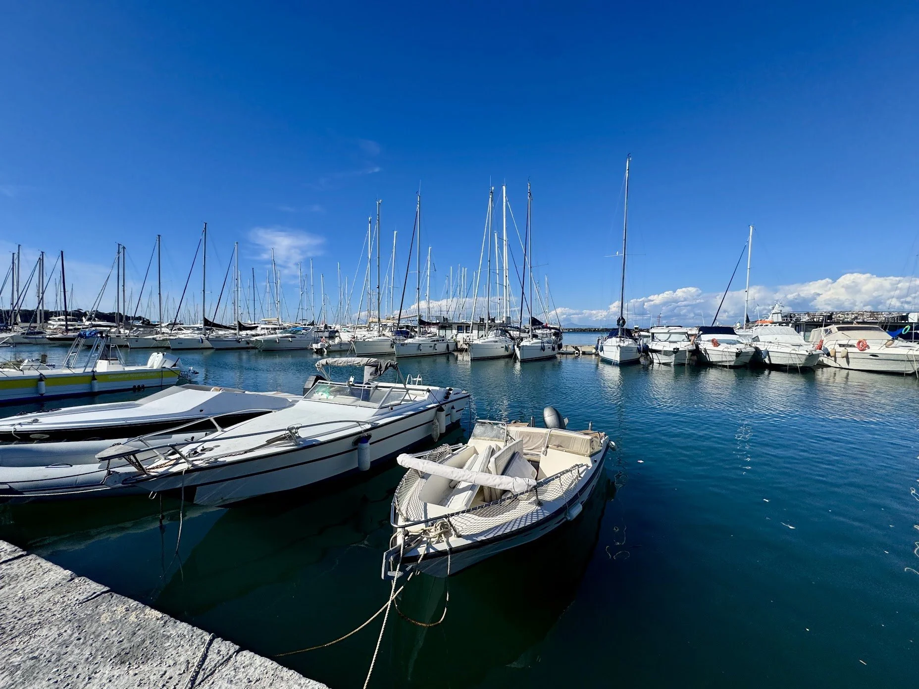 A marina filled with various boats and yachts docked in calm water under a clear blue sky with some clouds in the background.