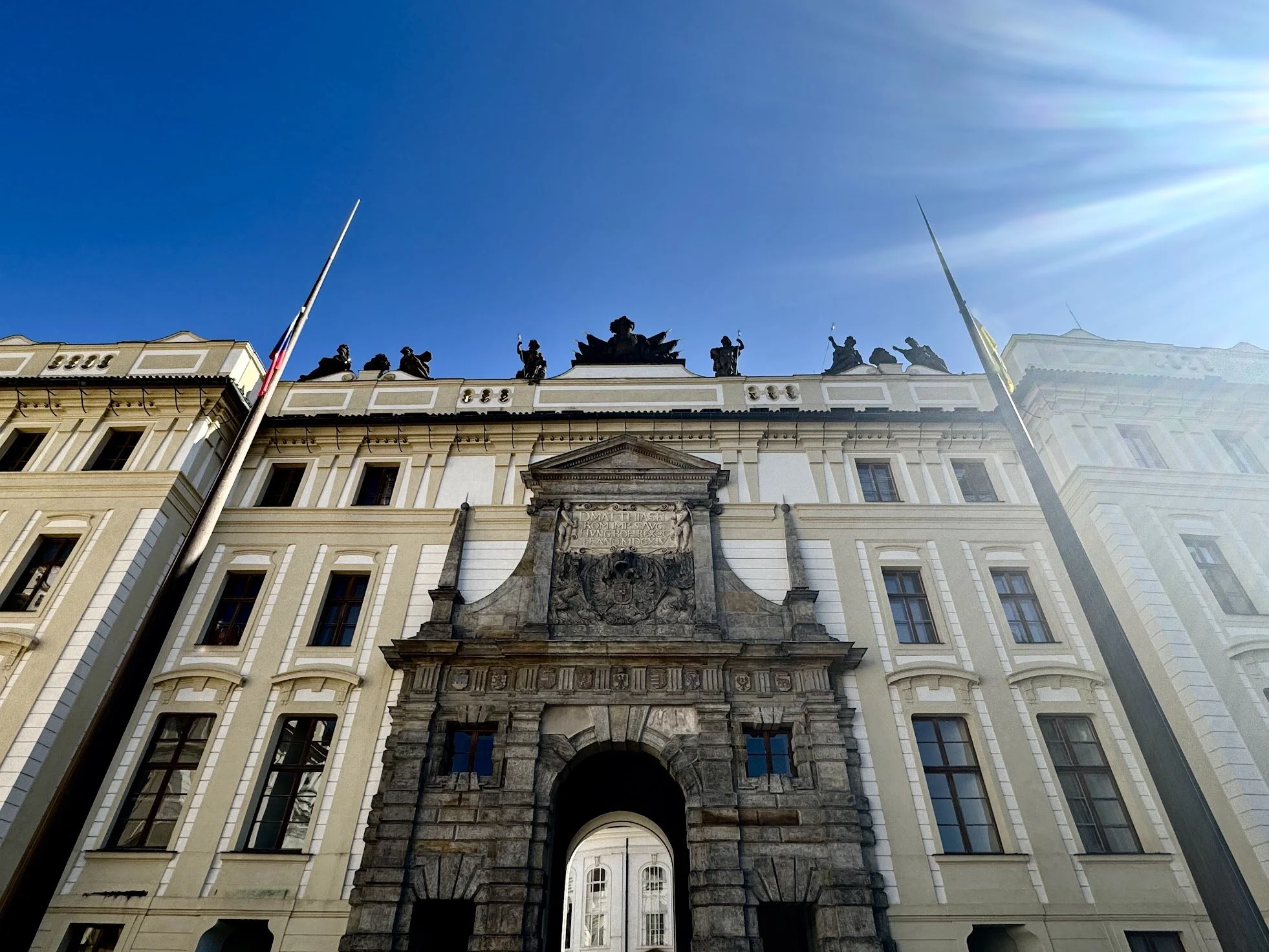Looking up at a historic government building with a large coat of arms above the main entrance, topped with statues holding flags, under a clear blue sky.