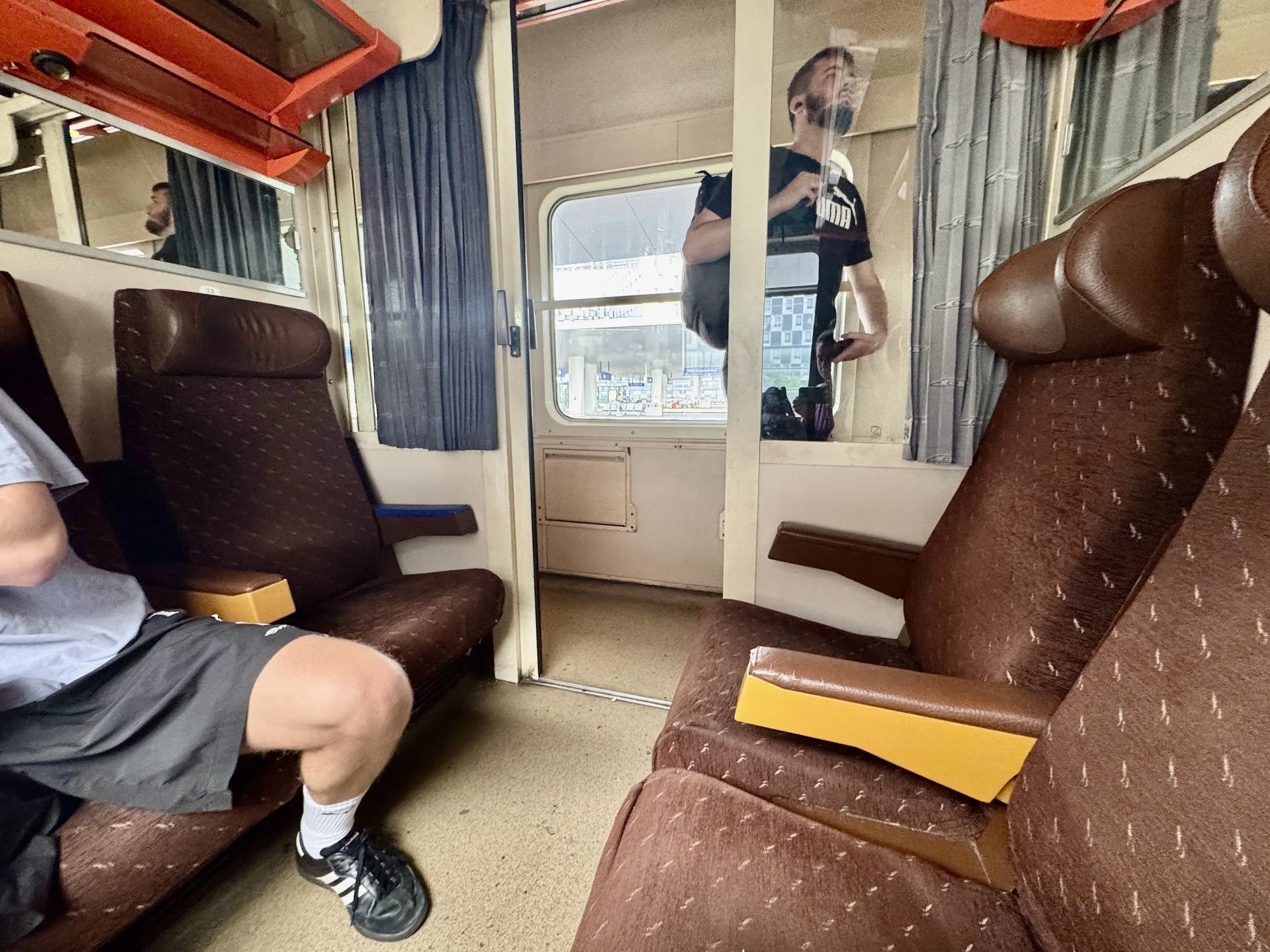 Interior of a train with brown fabric seats, a passenger wearing gray shorts and a gray shirt sitting on the left, and a man in black T-shirt with a backpack and face mask standing near the window. The window shows a train platform outside.